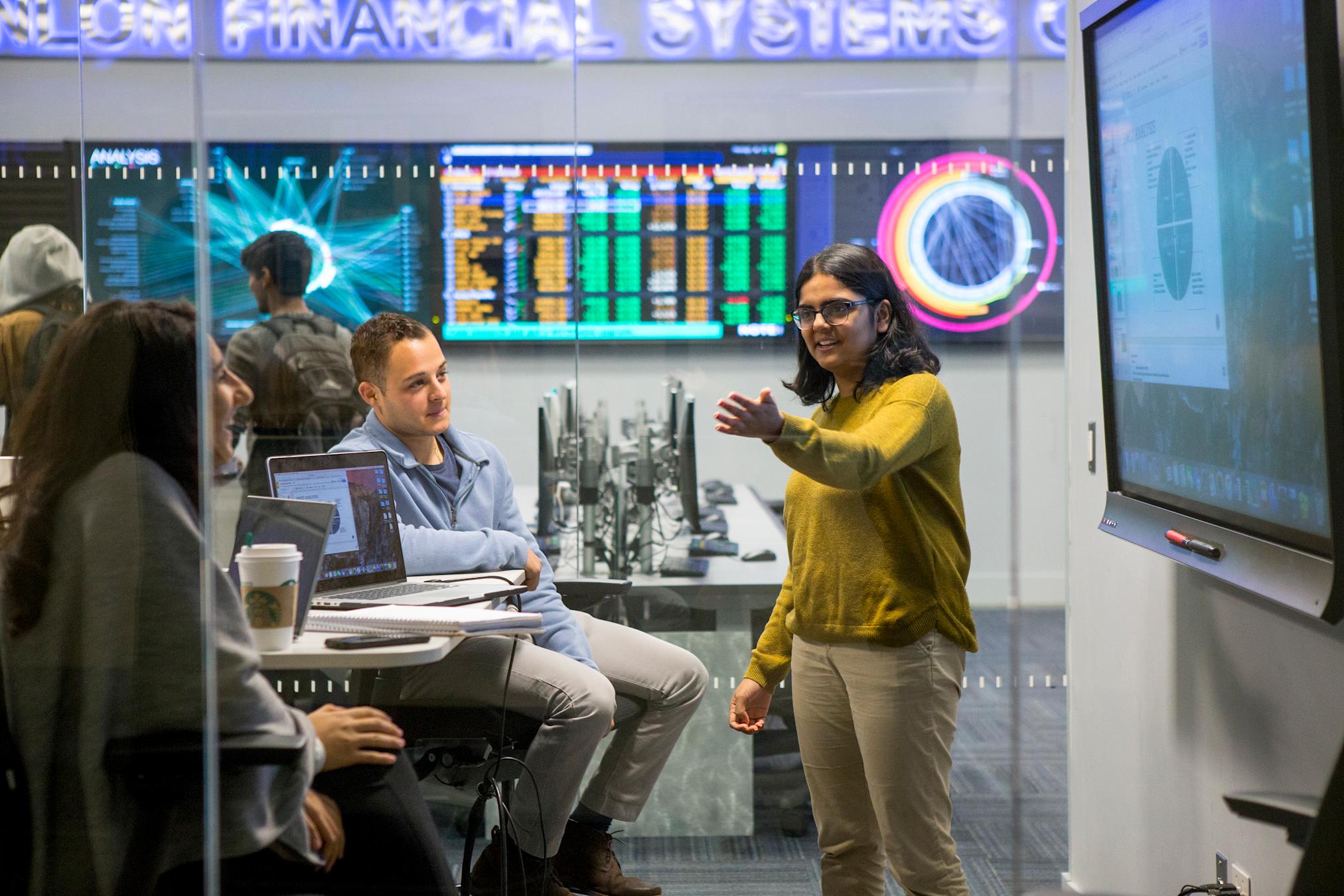 Student gestures to other students in front of monitors displaying data