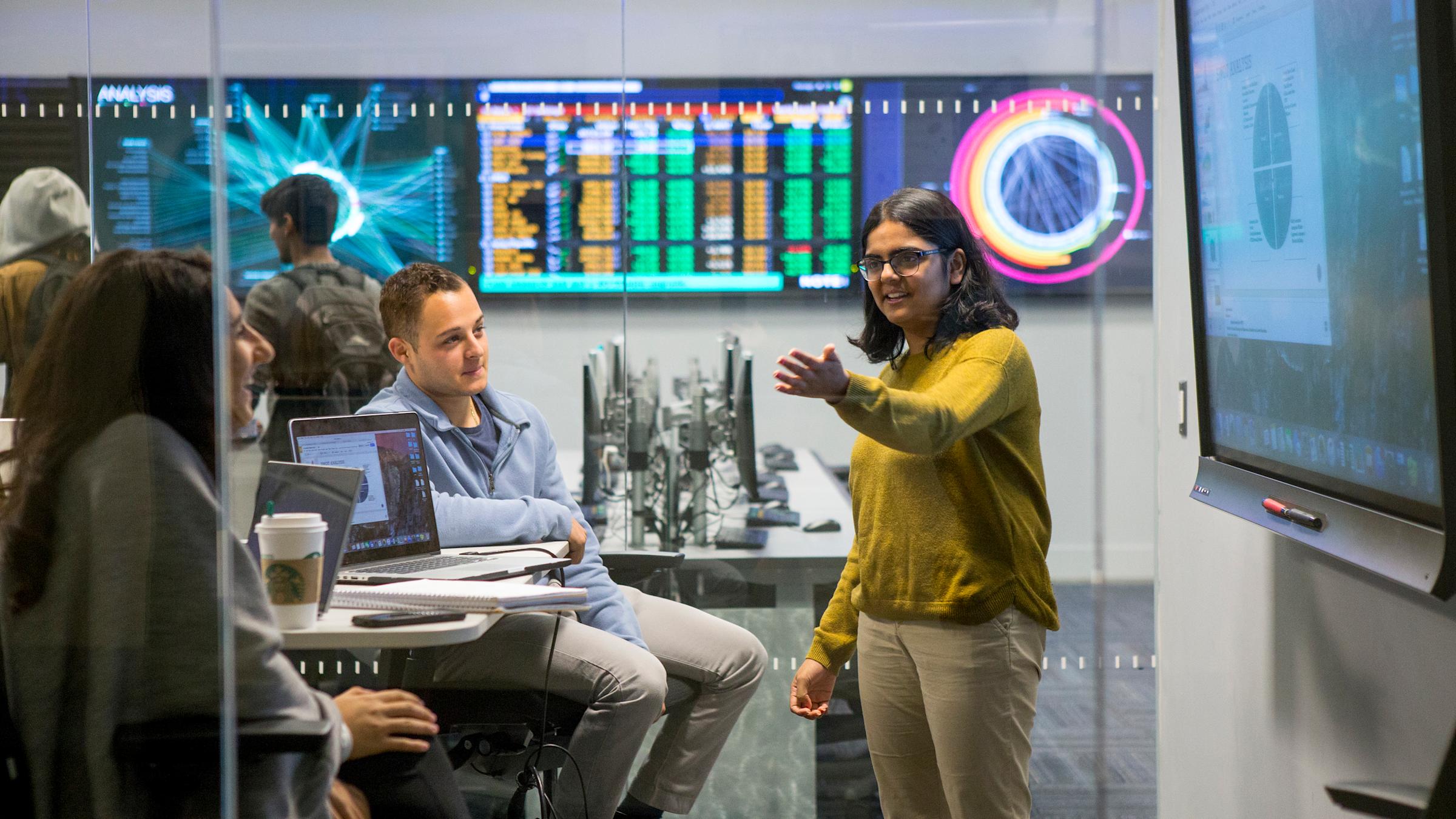Student gestures to other students in front of monitors displaying data