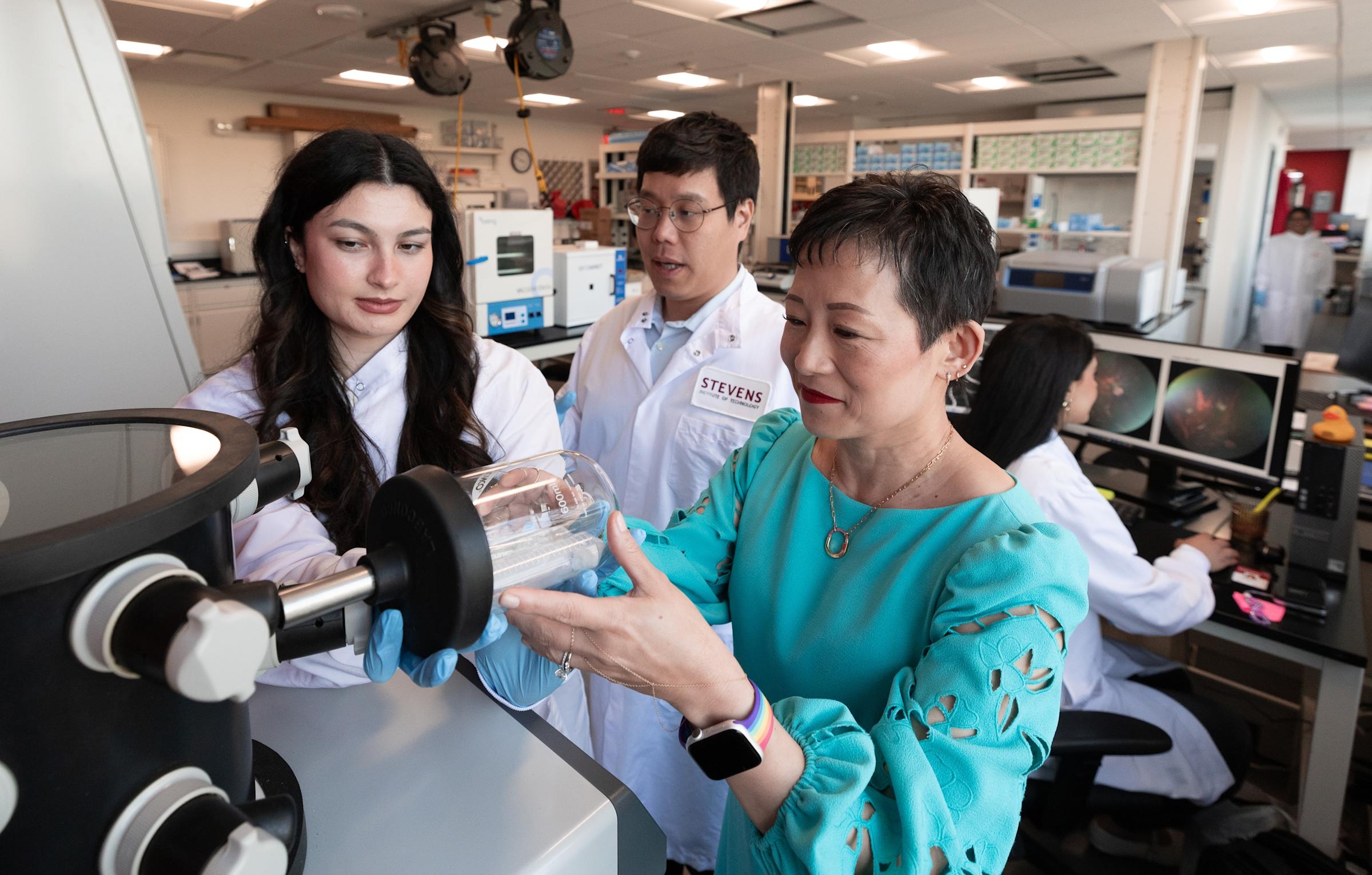 Jennifer Kang-Mieler in her lab demonstrating equipment for two students wearing lab coats.