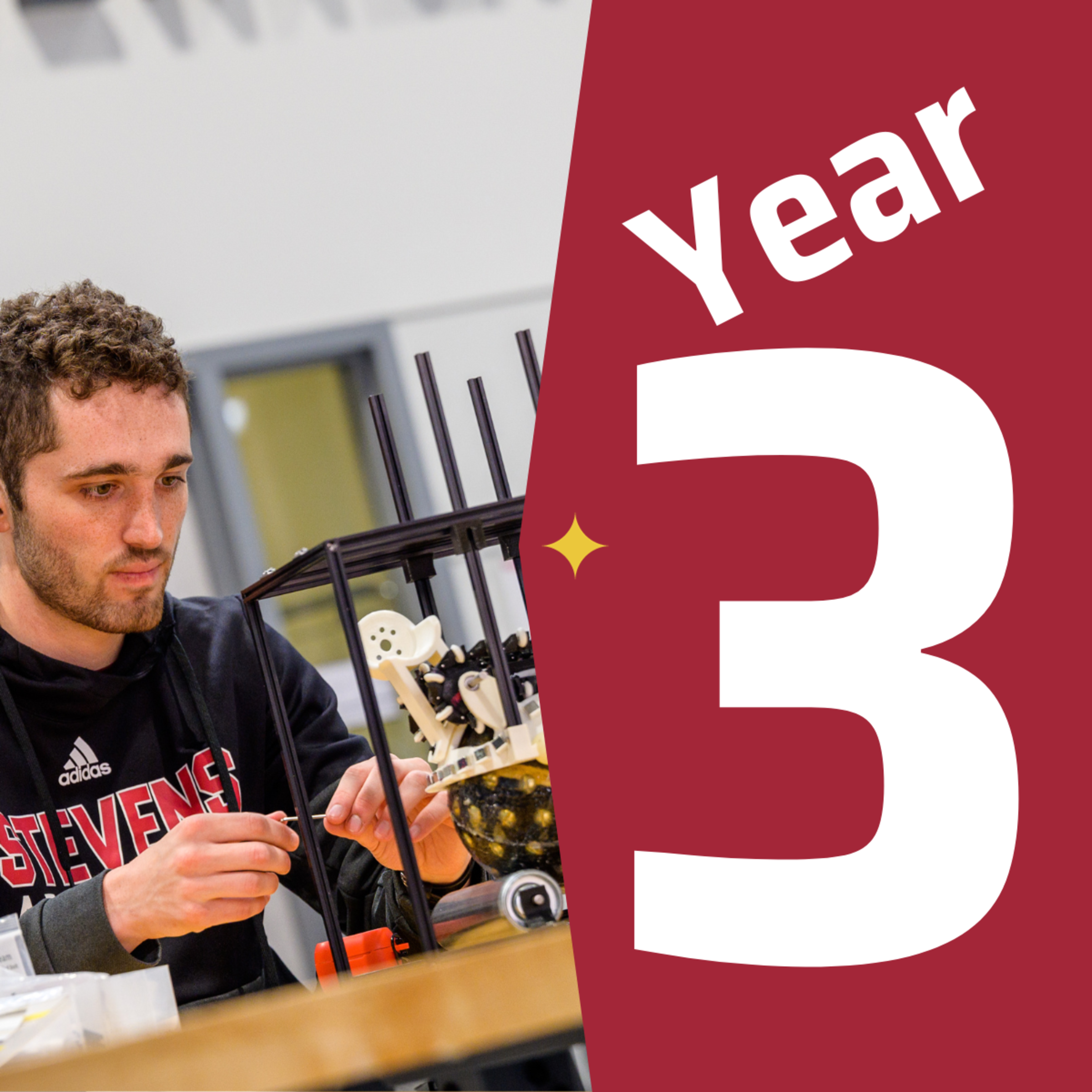 A male student wearing a Stevens shirt working on an engineering project next to a graphic that says year three.
