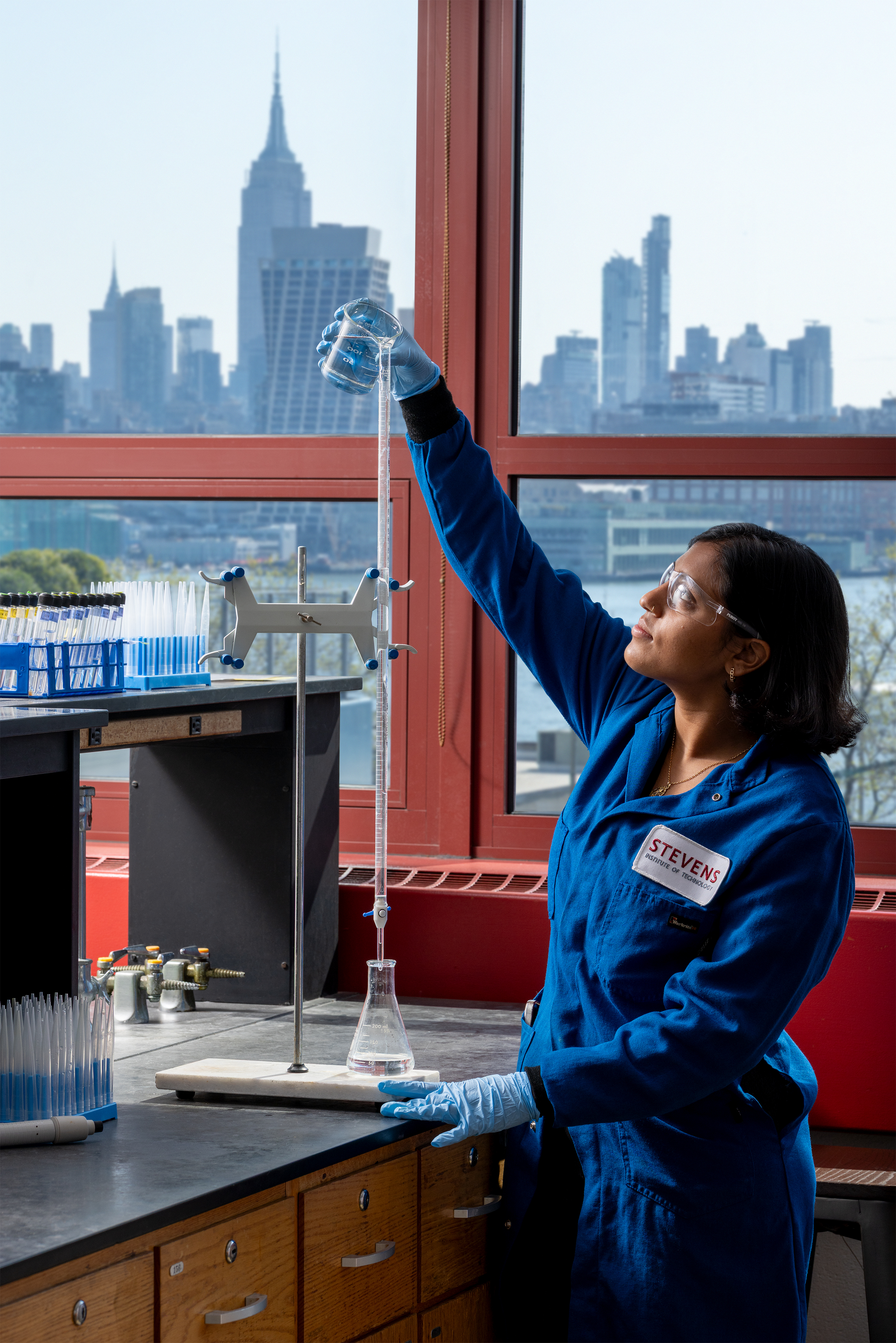 A female student in a blue jumpsuit raising a glass in a lab