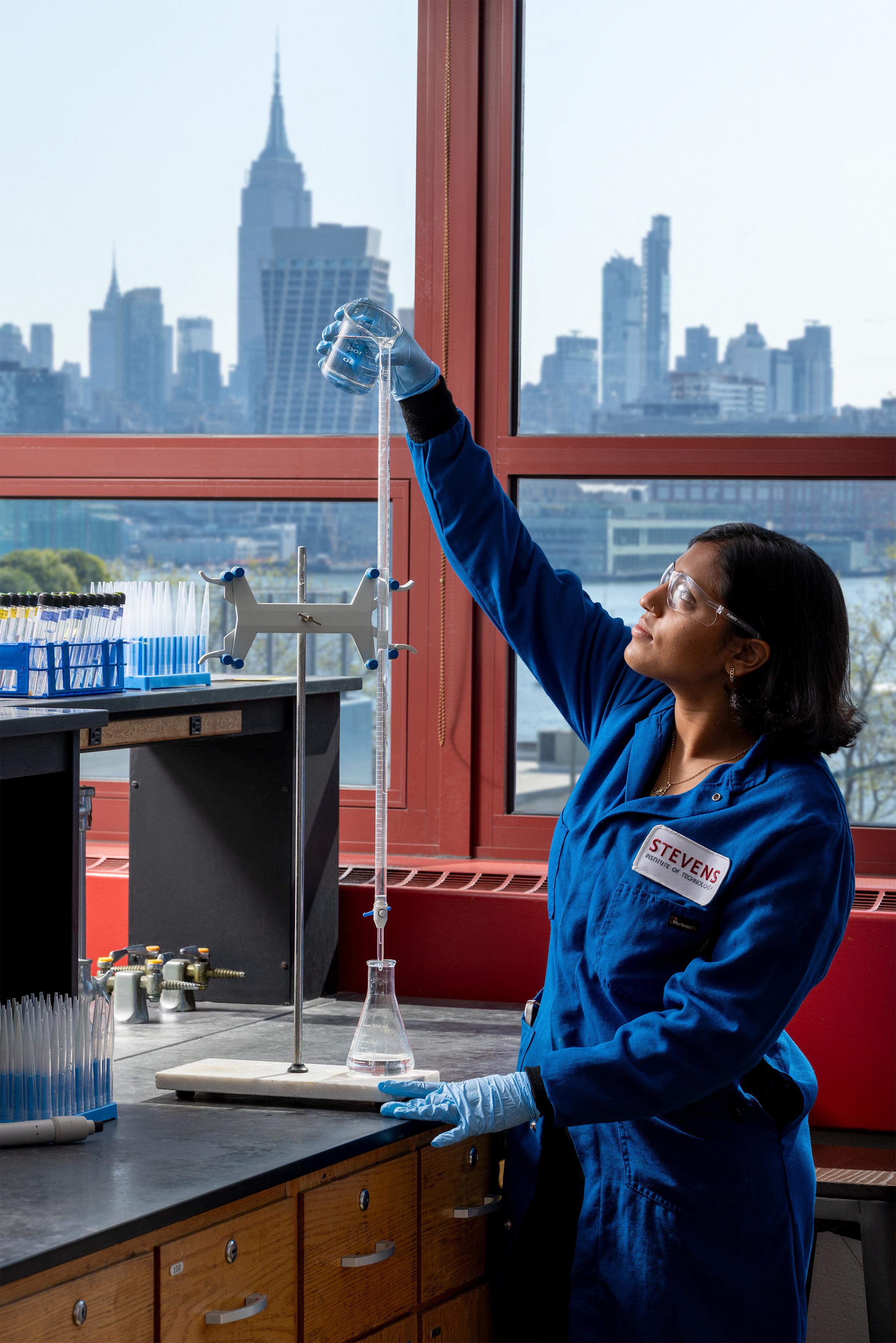 A female student in a blue jumpsuit raising a glass in a lab