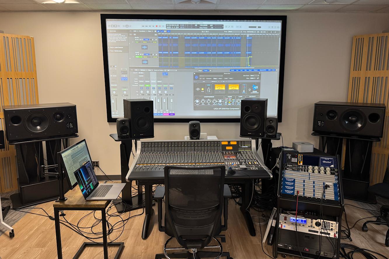 Large mixing board with speakers on either side and a wall monitor showing audio settings.