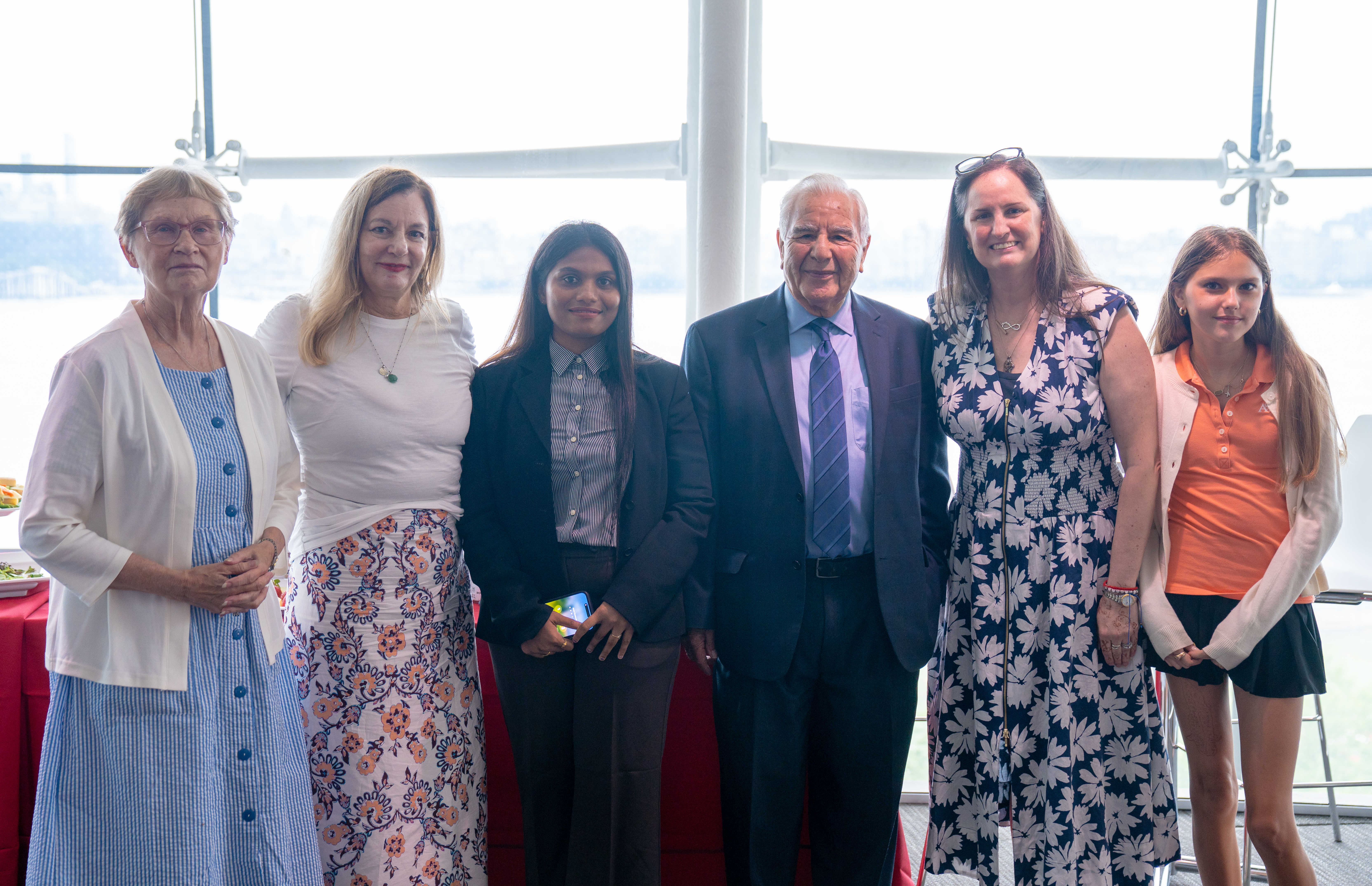 Six people pose together in a bright room with large windows overlooking a waterway. The group includes a woman in a blue dress, a woman in a floral skirt and white top, a woman in business attire, a man in a suit and tie, and two women in summer dresses.
