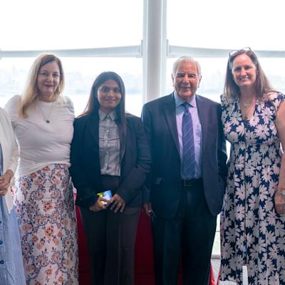 Six people pose together in a bright room with large windows overlooking a waterway. The group includes a woman in a blue dress, a woman in a floral skirt and white top, a woman in business attire, a man in a suit and tie, and two women in summer dresses.