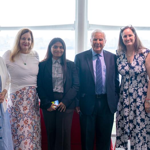 Six people pose together in a bright room with large windows overlooking a waterway. The group includes a woman in a blue dress, a woman in a floral skirt and white top, a woman in business attire, a man in a suit and tie, and two women in summer dresses.