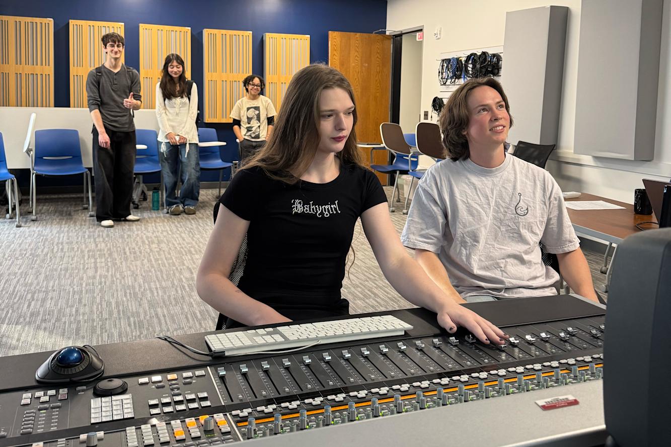 Two students working at a mixing board in a large room with other students.