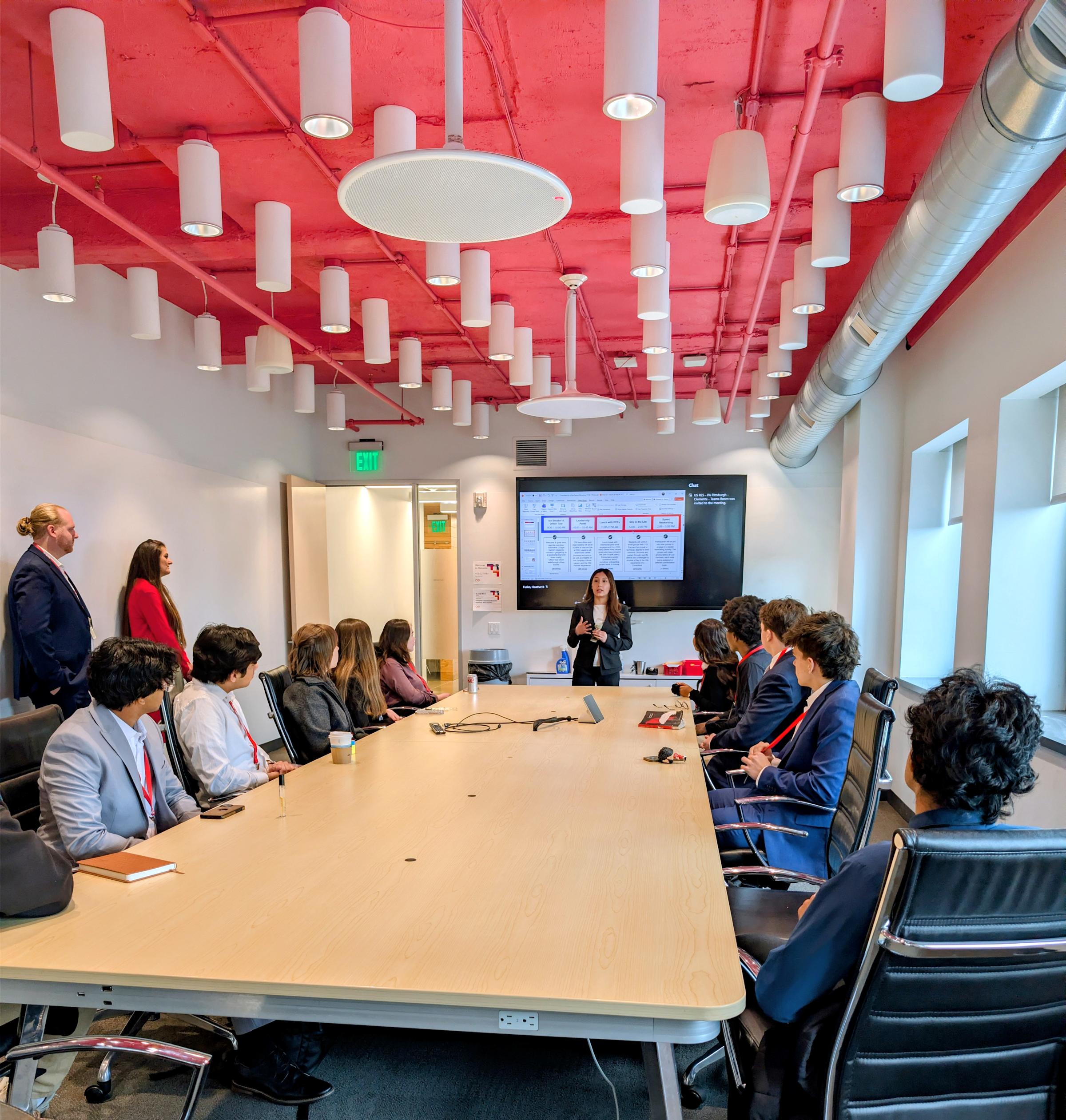 Students wearing business attire and red lanyards sit around a long conference table in a modern room with exposed red ceiling, white cylindrical pendant lights, and large windows. A female presenter stands at the front gesturing toward a large screen.