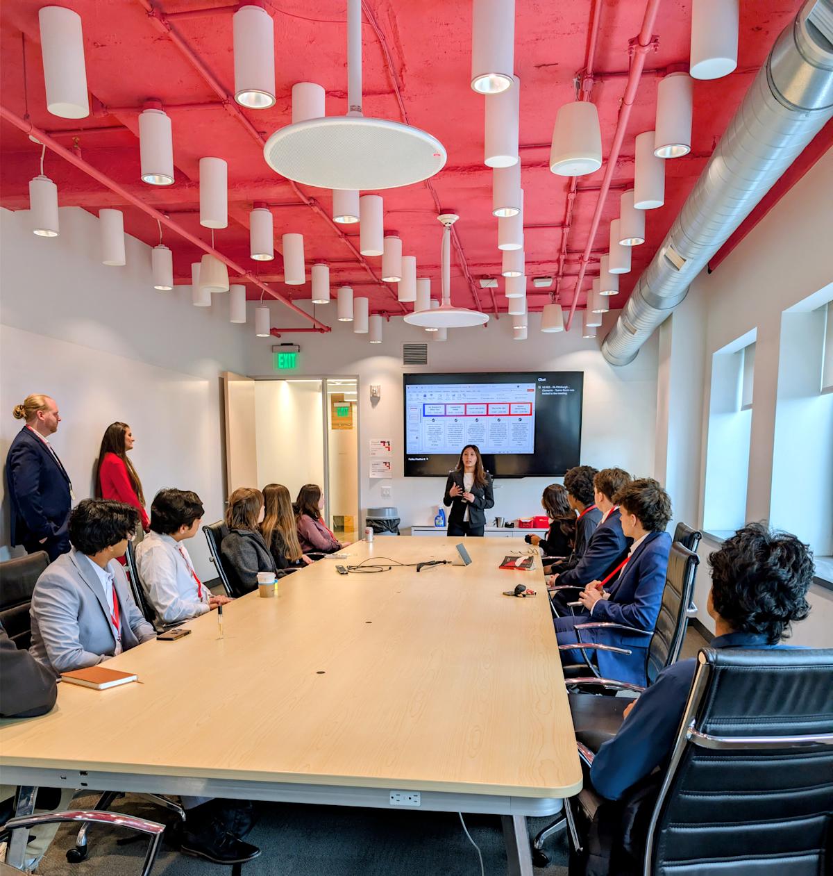 Students wearing business attire and red lanyards sit around a long conference table in a modern room with exposed red ceiling, white cylindrical pendant lights, and large windows. A female presenter stands at the front gesturing toward a large screen.