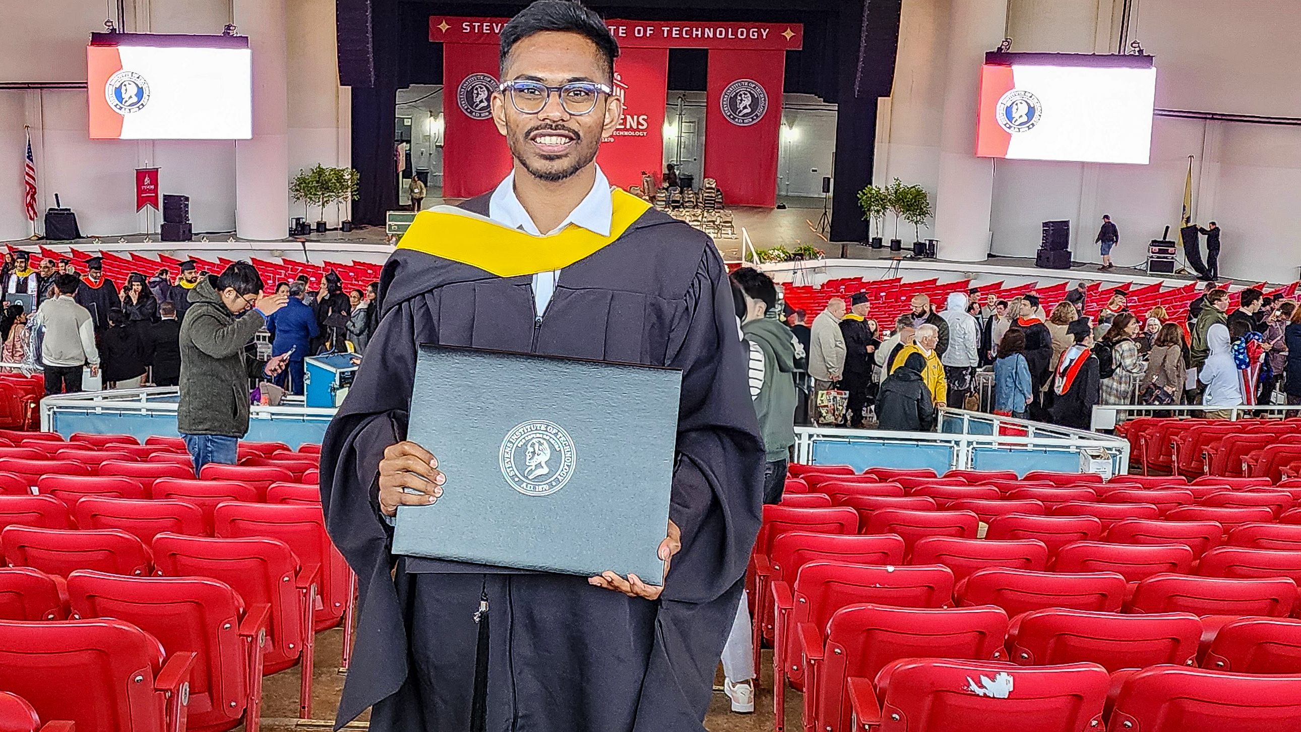 Aaryan Yekkisetty holds up his diploma wearing his black gown at his Stevens Institute of Technology graduation.