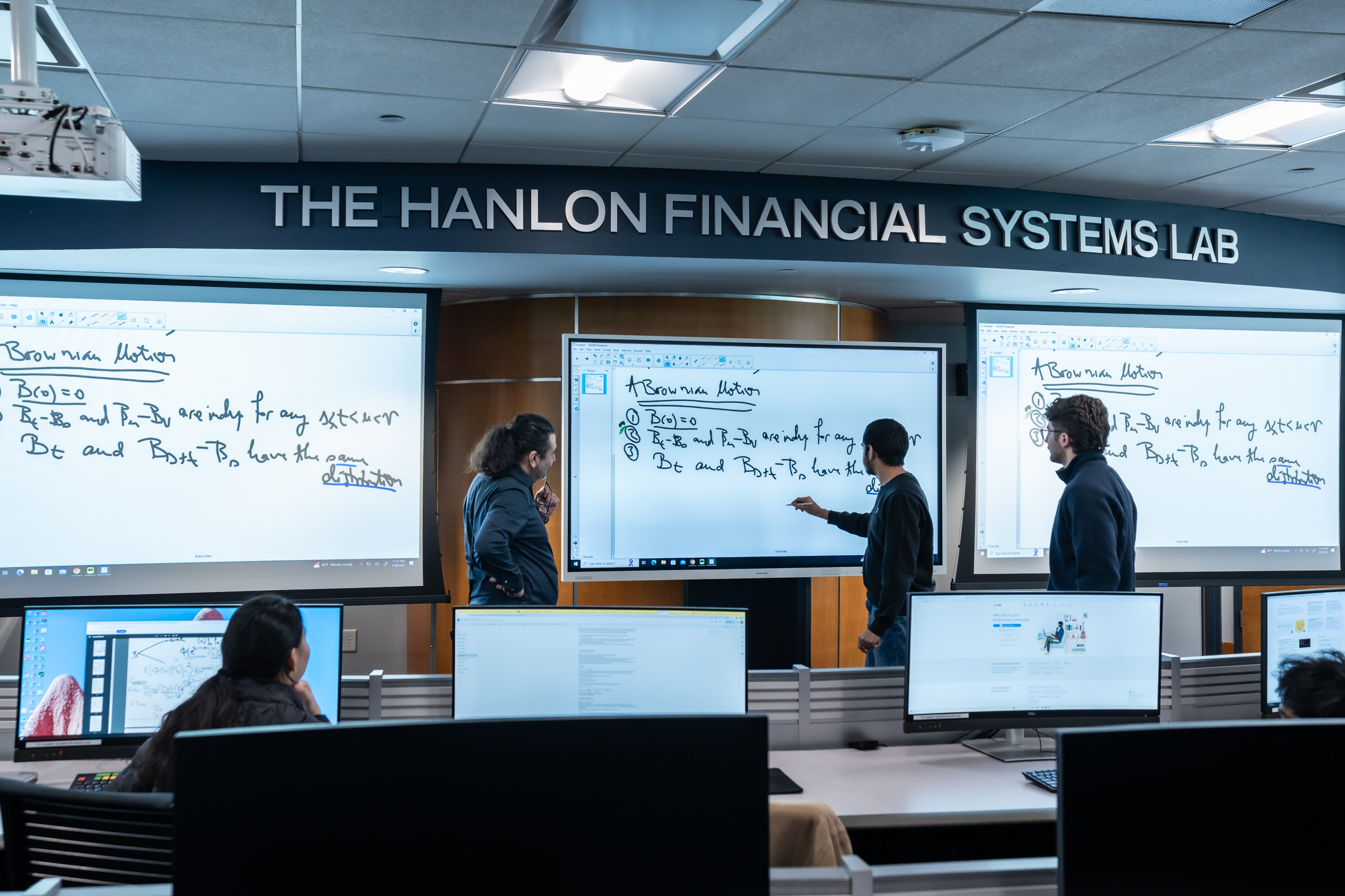 Students collaborate in The Hanlon Financial Systems Lab, working at computer workstations while three stand at large digital whiteboards displaying mathematical equations. The modern trading lab features multiple monitors and tiered seating.