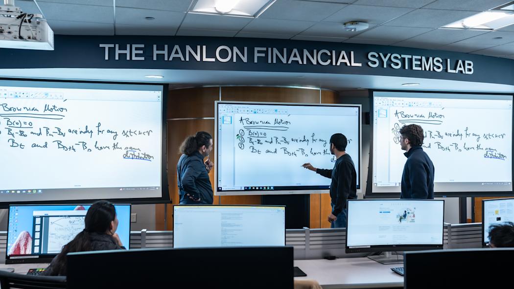 Students collaborate in The Hanlon Financial Systems Lab, working at computer workstations while three stand at large digital whiteboards displaying mathematical equations. The modern trading lab features multiple monitors and tiered seating.