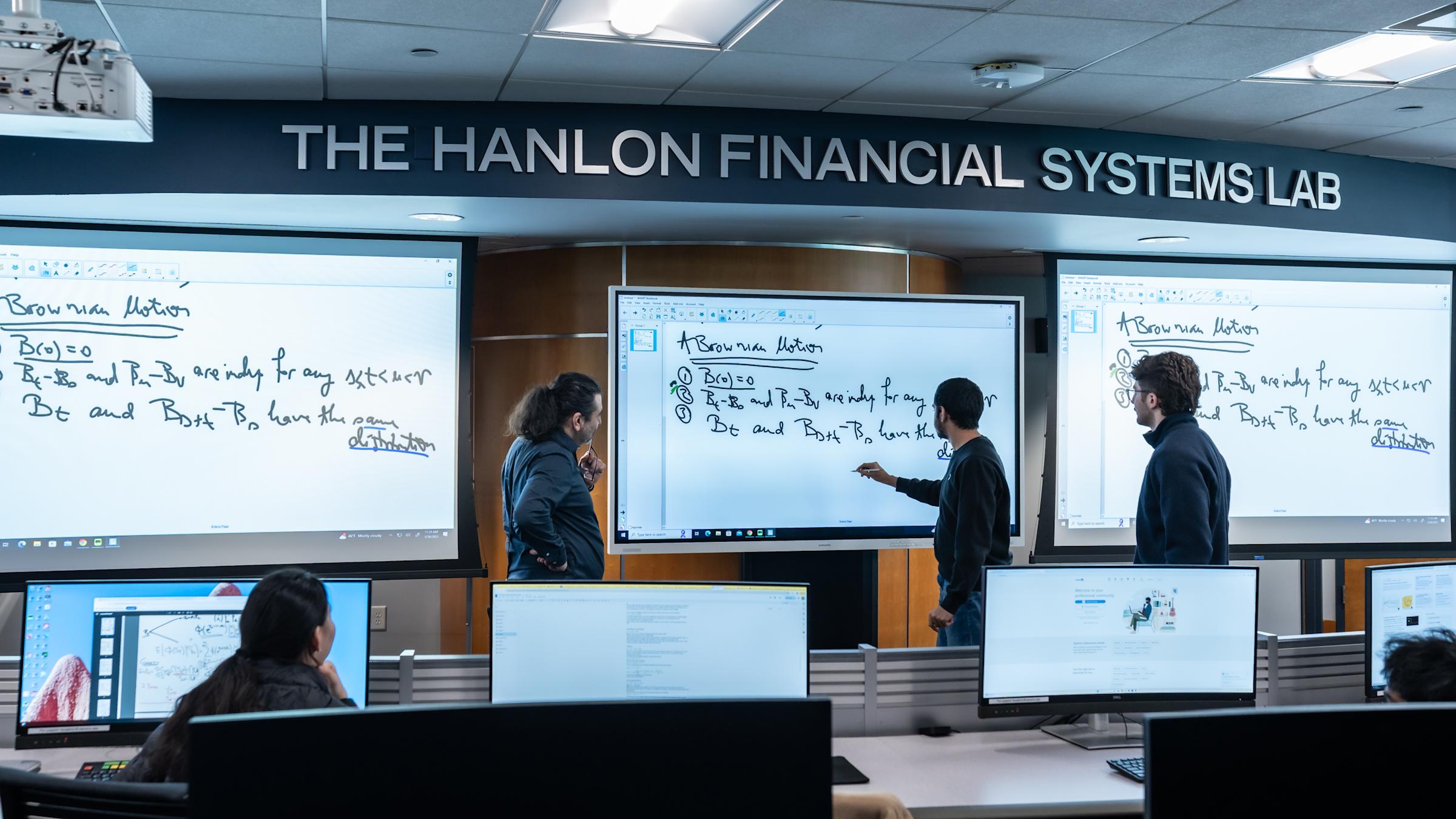 Students collaborate in The Hanlon Financial Systems Lab, working at computer workstations while three stand at large digital whiteboards displaying mathematical equations. The modern trading lab features multiple monitors and tiered seating.