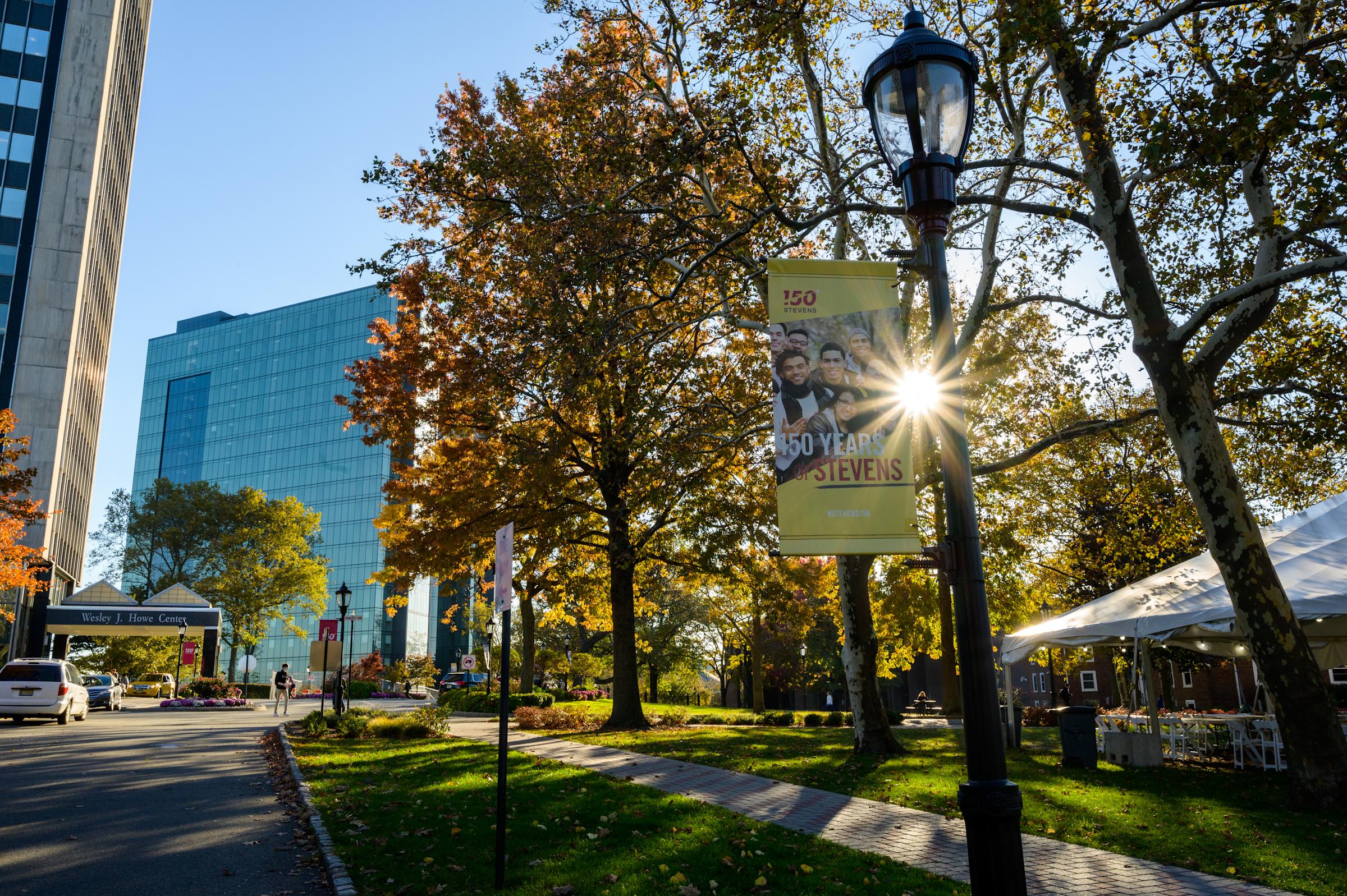 Campus image with trees and University Center in background. Lightpole banner highlighting 150 years of Stevens.