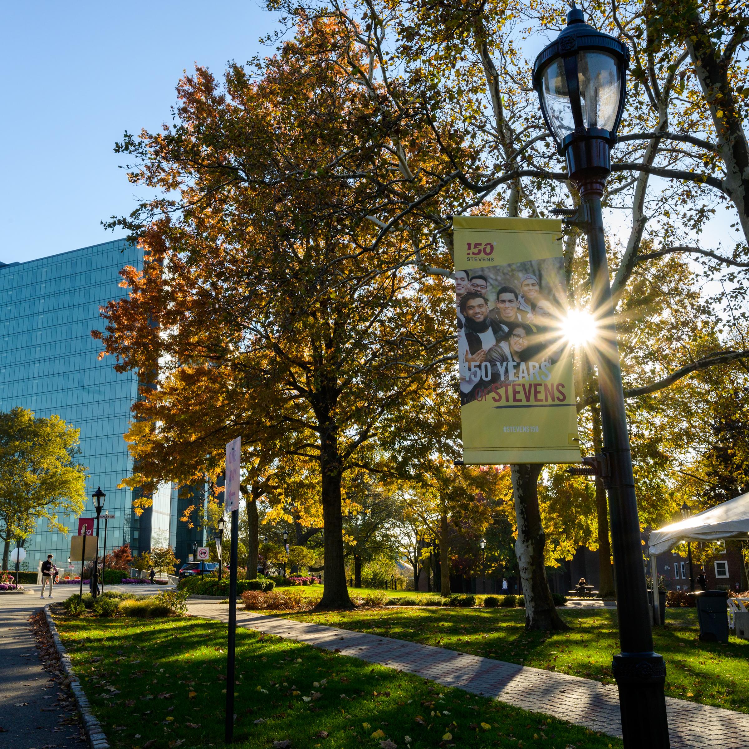 Campus image with trees and University Center in background. Lightpole banner highlighting 150 years of Stevens.