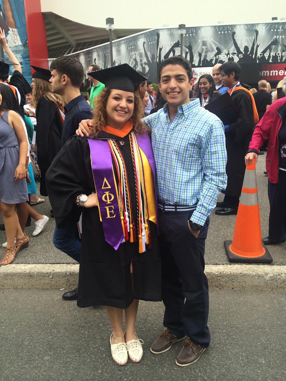 A woman in a black graduation cap and gown with a purple Greek-letter stole and multiple honor cords stands smiling next to a man in a green and blue plaid shirt, his arm around her shoulder, on a paved area near a stadium.