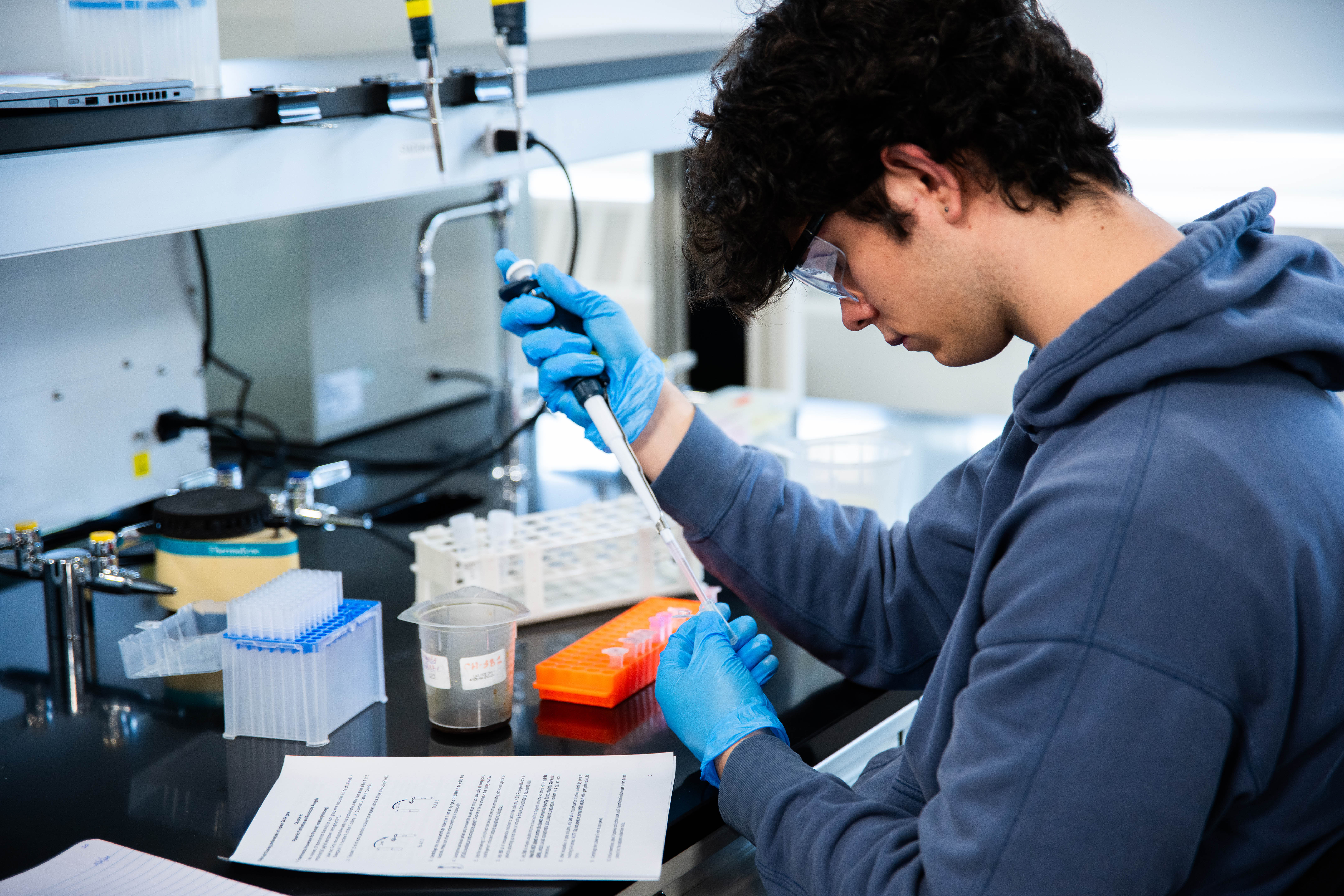 A chemistry and chemical biology student with gloves handles an injection into a glass tube.