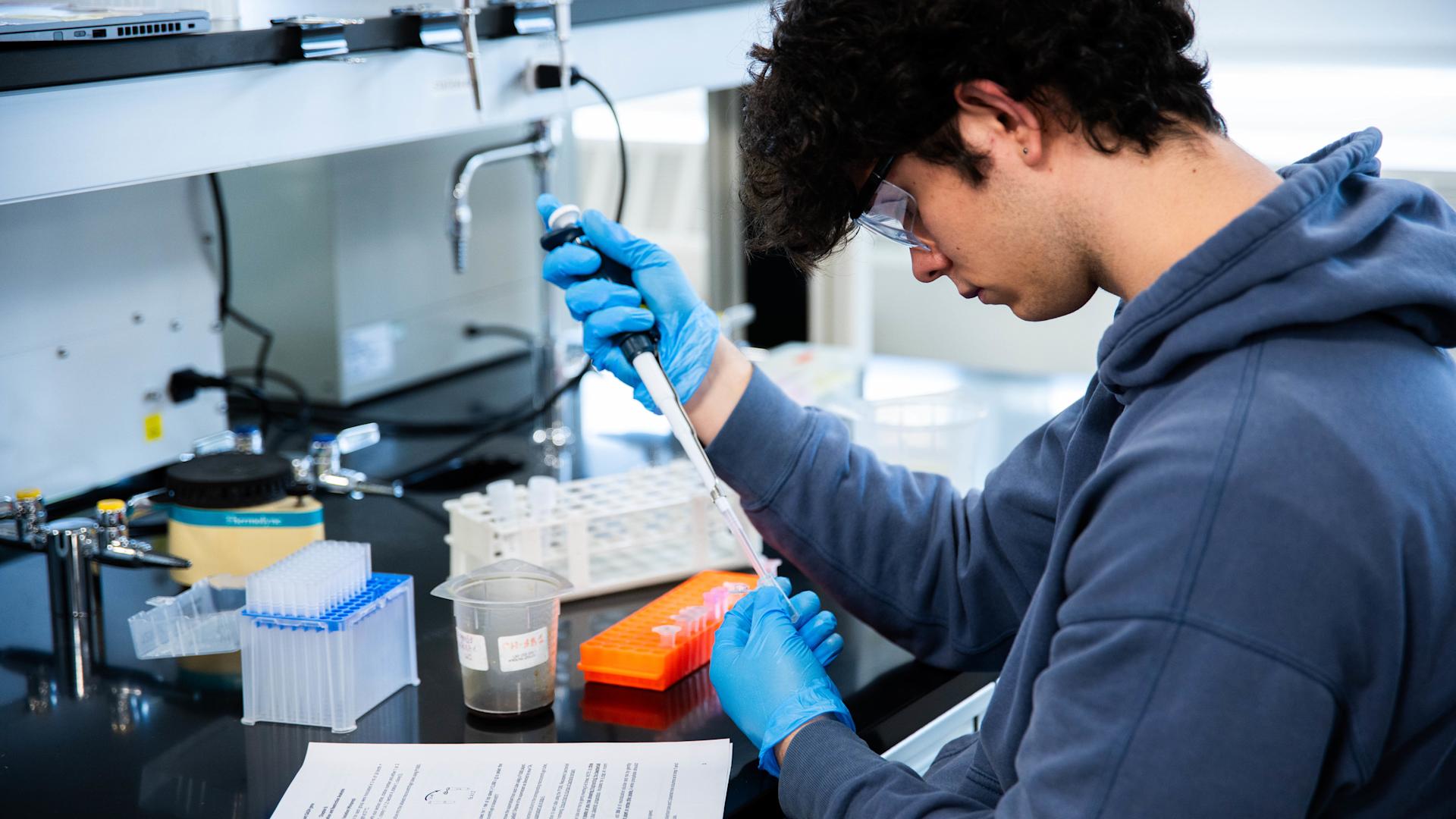 A chemistry and chemical biology student with gloves handles an injection into a glass tube.