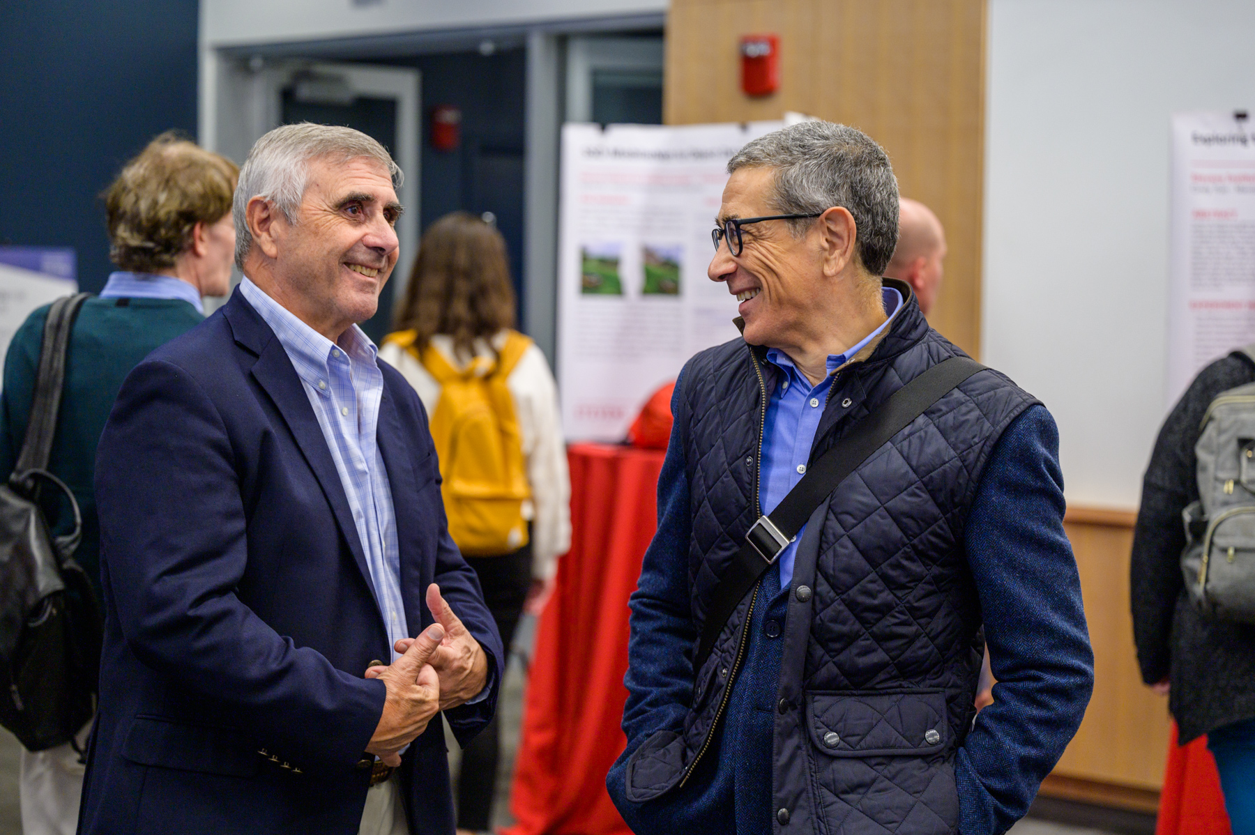 Anthony Barrese and Carlo Lipizzi smiling and talking with each other at the SIAI Relaunch Event, with posters and other attendees visible in the background.