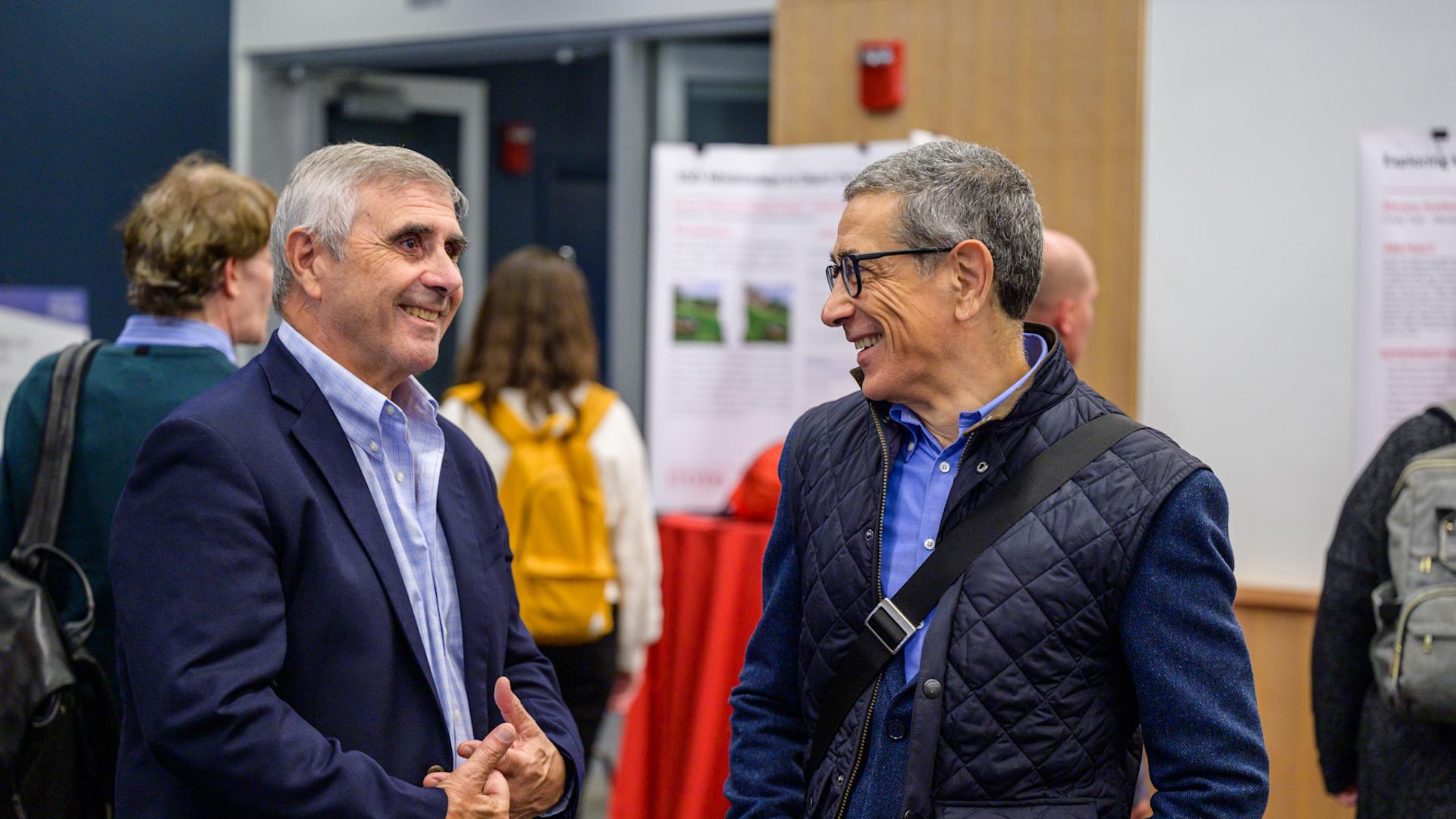 Anthony Barrese and Carlo Lipizzi smiling and talking with each other at the SIAI Relaunch Event, with posters and other attendees visible in the background.