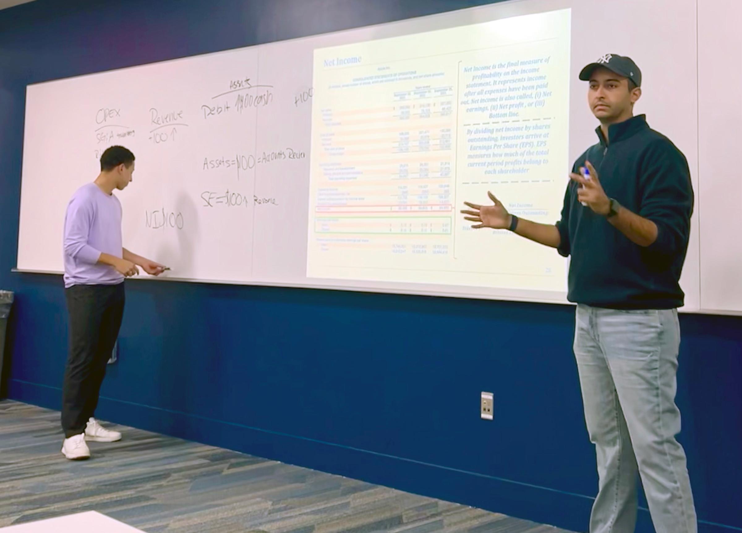 Two people presenting financial concepts in a classroom with whiteboard
