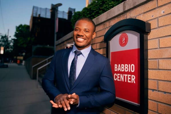 A photograph of Christopher Mbonu standing in front of a Stevens school building sign that reads Babbio Center.