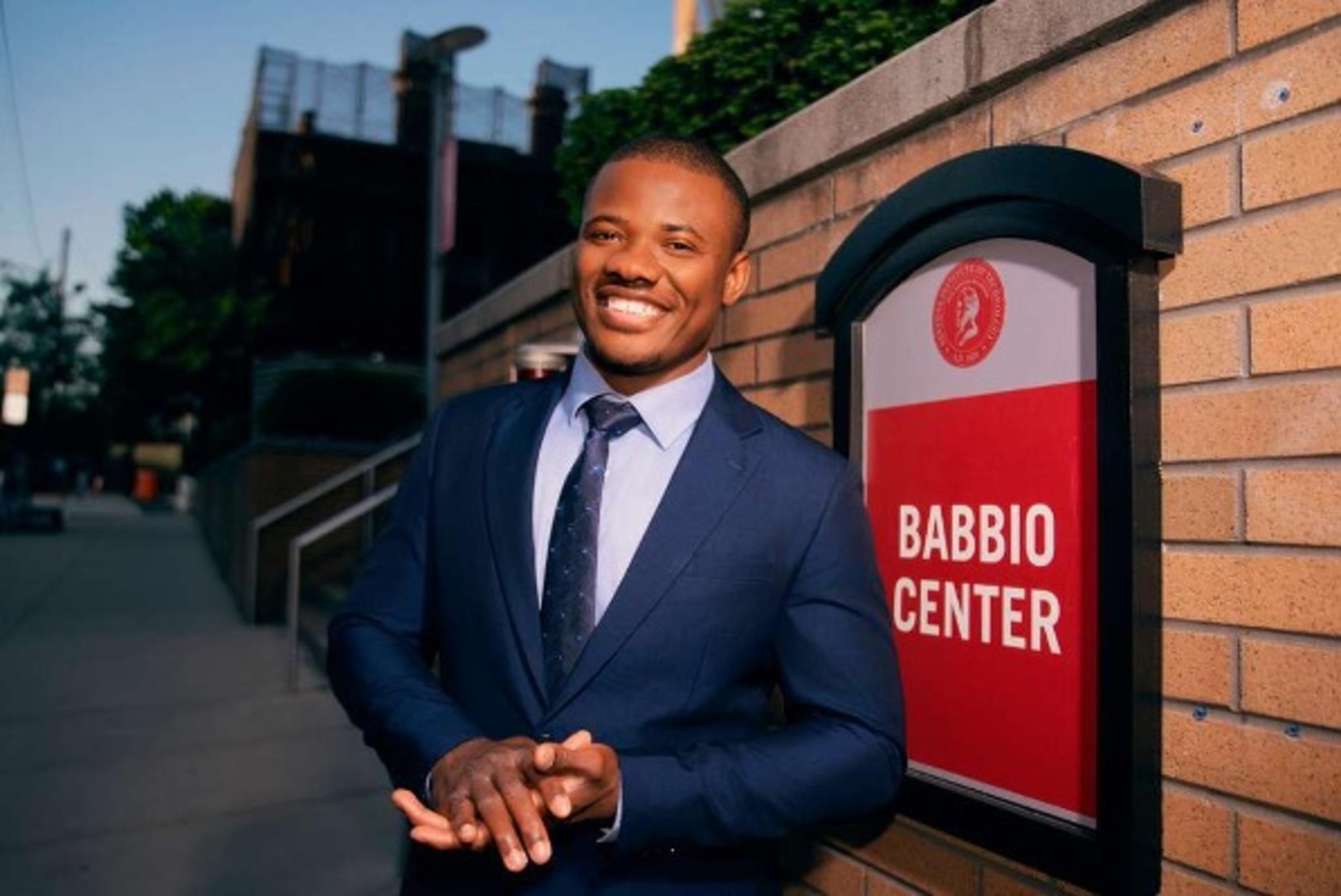 A photograph of Christopher Mbonu standing in front of a Stevens school building sign that reads Babbio Center.