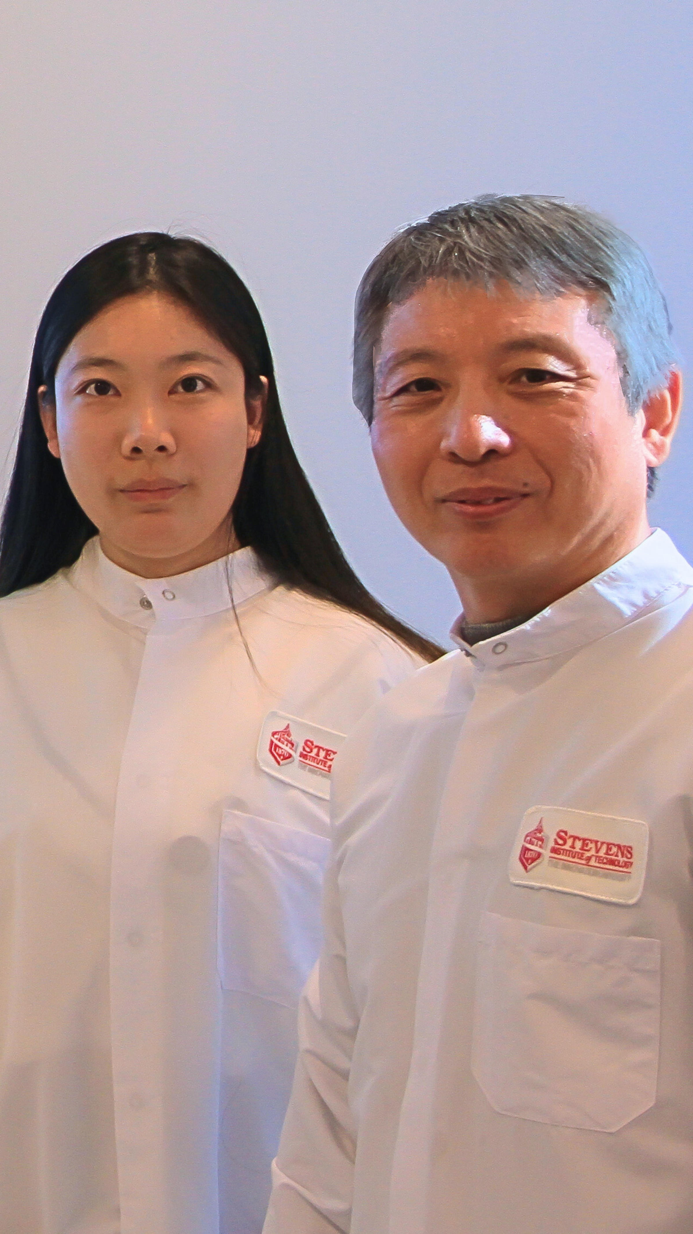 A woman with black hair on the left with a man with gray hair, both wearing lab coats with the Stevens Institute of Technology logo.