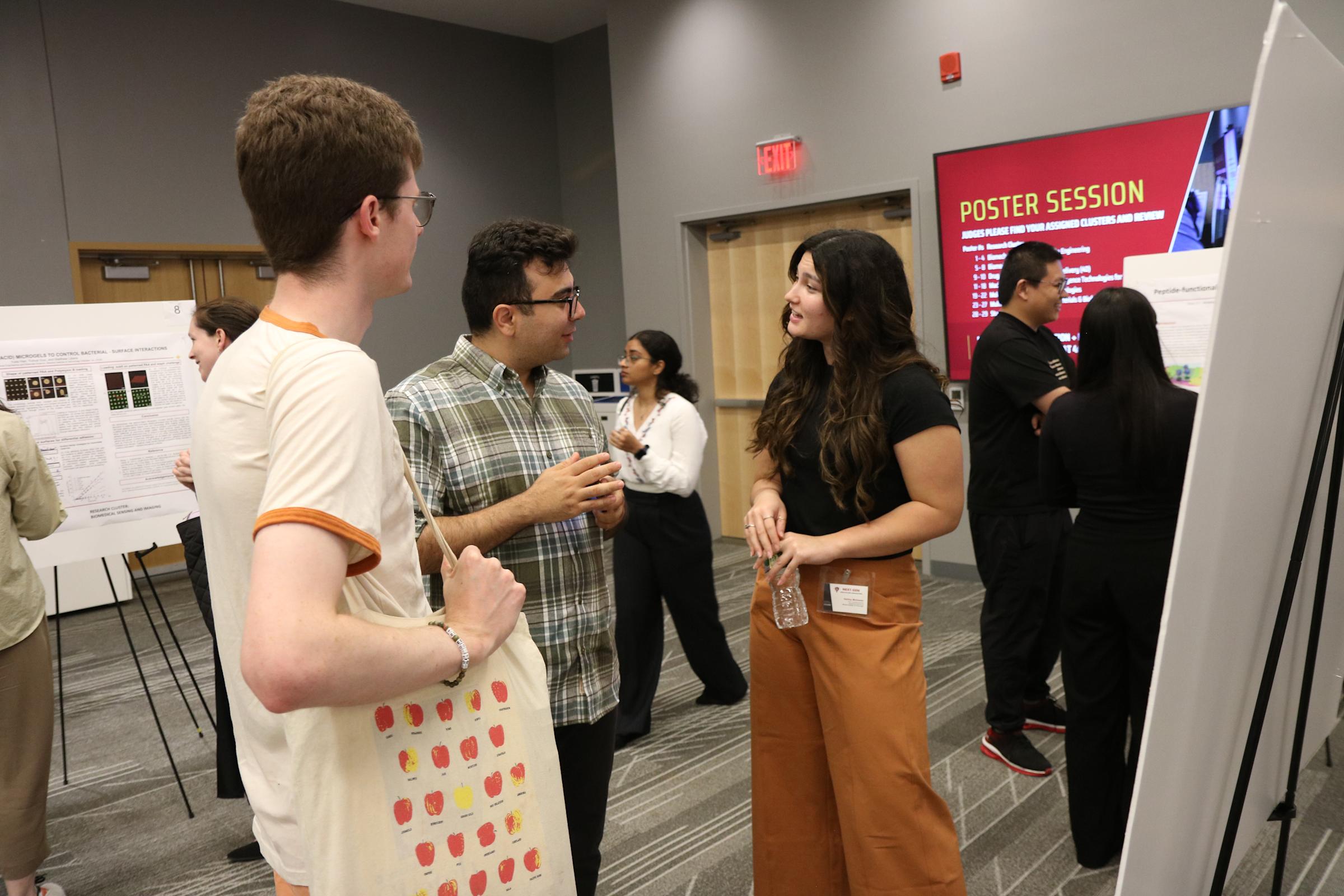 Students and research presenters interact in front of monitors and posters in a auditorium.