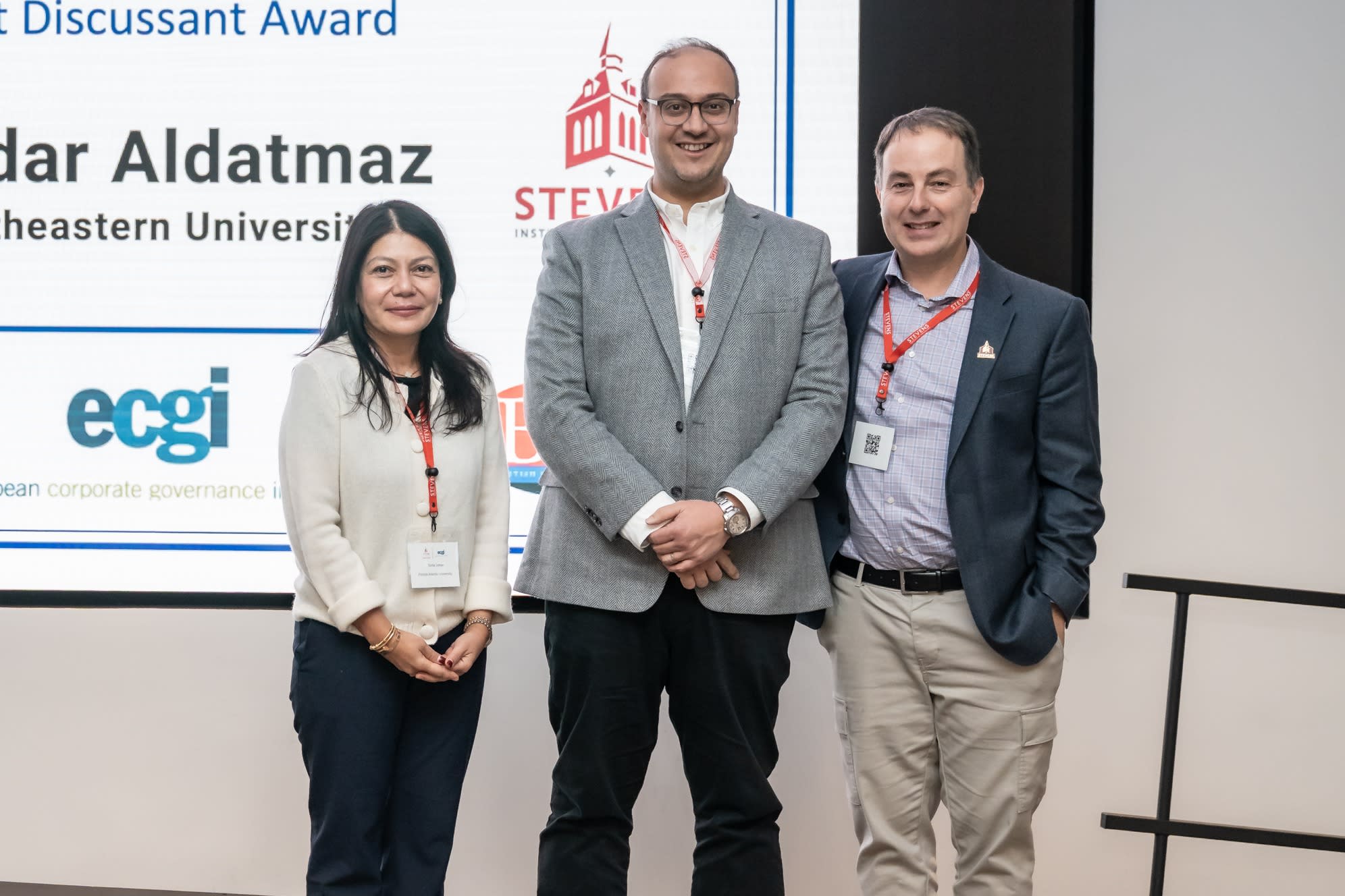Three people pose smiling in front of a large screen displaying "RCF-ECGI Corporate Finance and Governance 2025 Best Discussant Award" honoring Serdar Aldatmaz of Northeastern University.