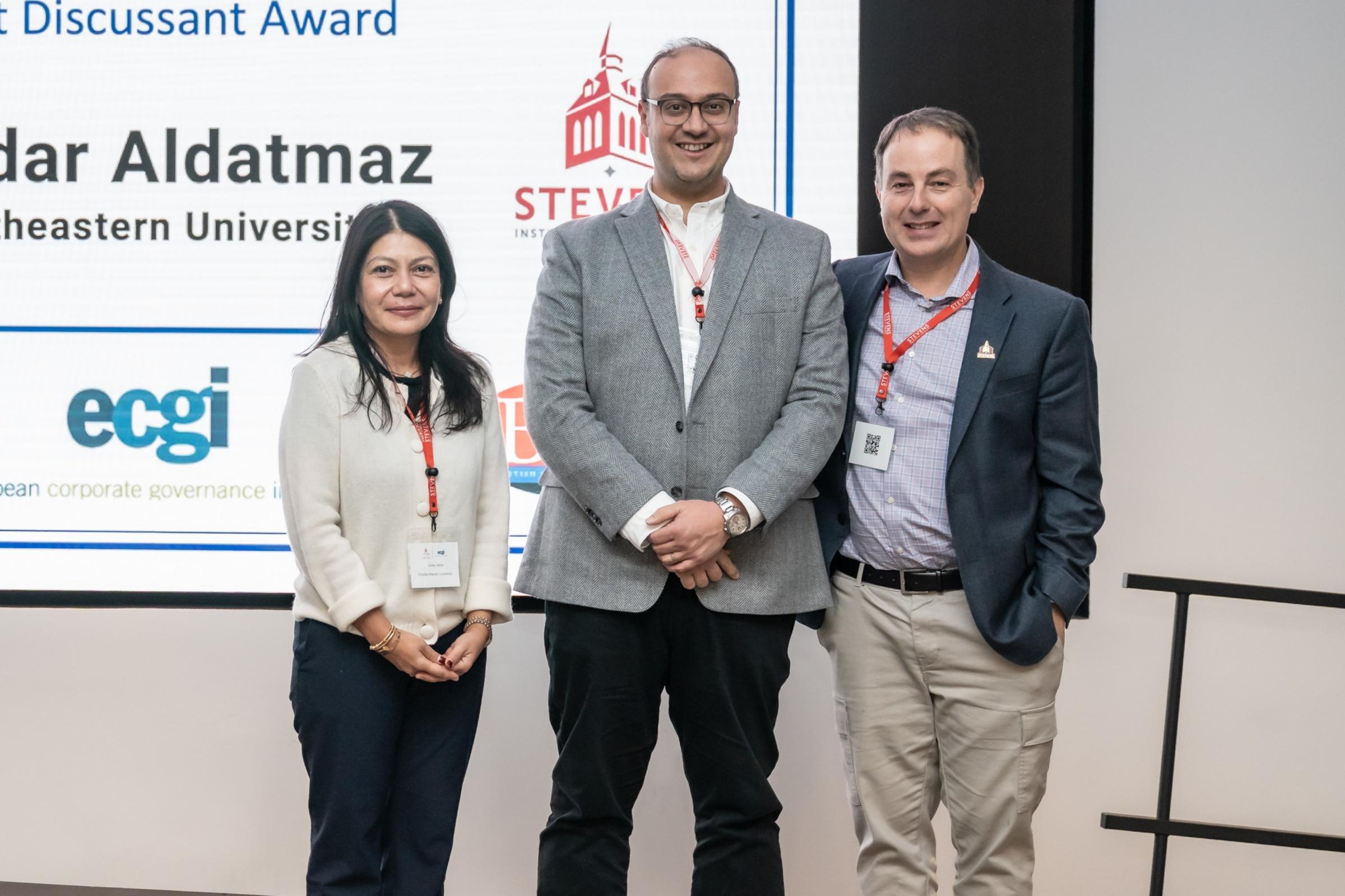 Three people pose smiling in front of a large screen displaying "RCF-ECGI Corporate Finance and Governance 2025 Best Discussant Award" honoring Serdar Aldatmaz of Northeastern University.