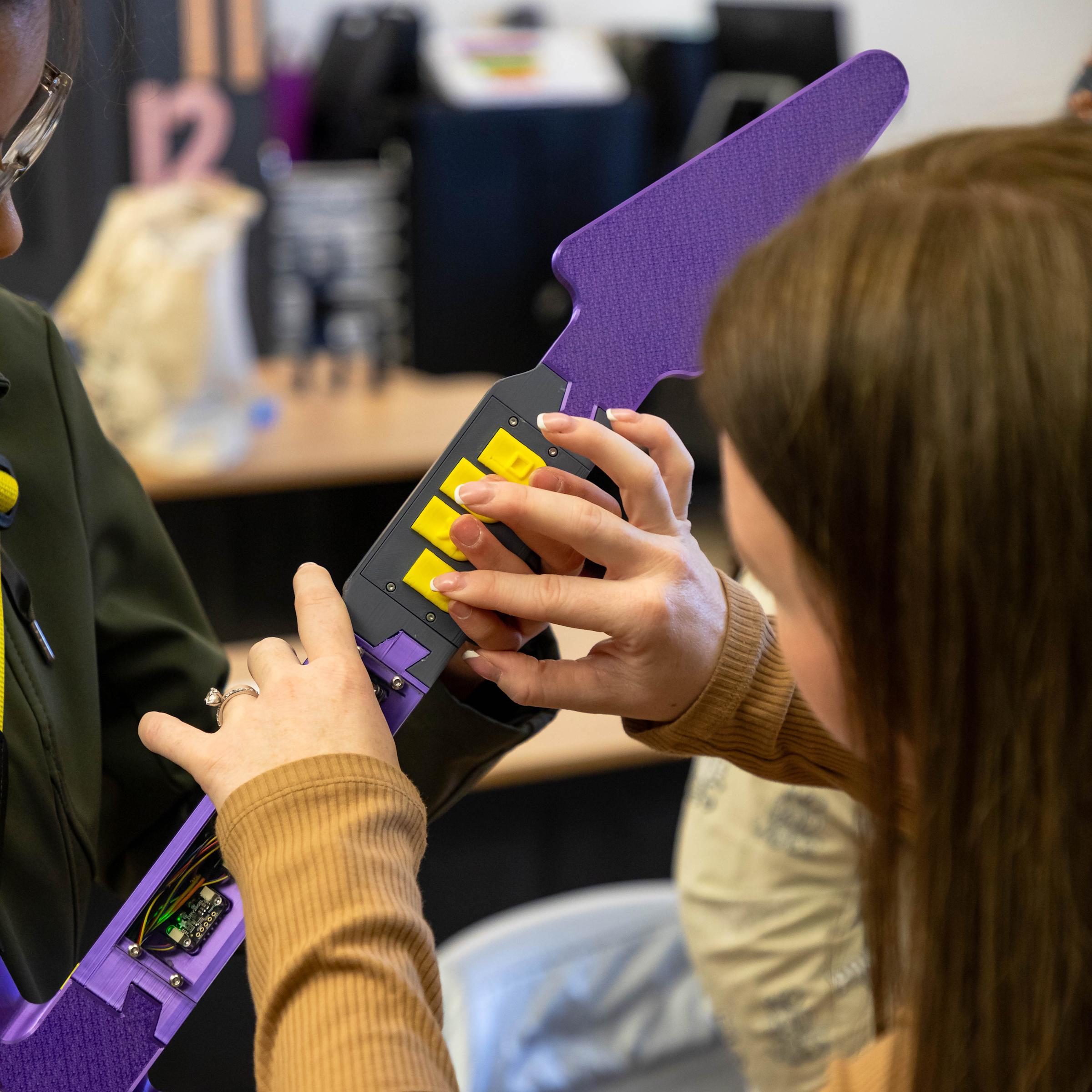 A student on the left plays a purple electric guitar and is guided by a teacher who helps her press the correct guitar buttons of the Haptic Hero game.
