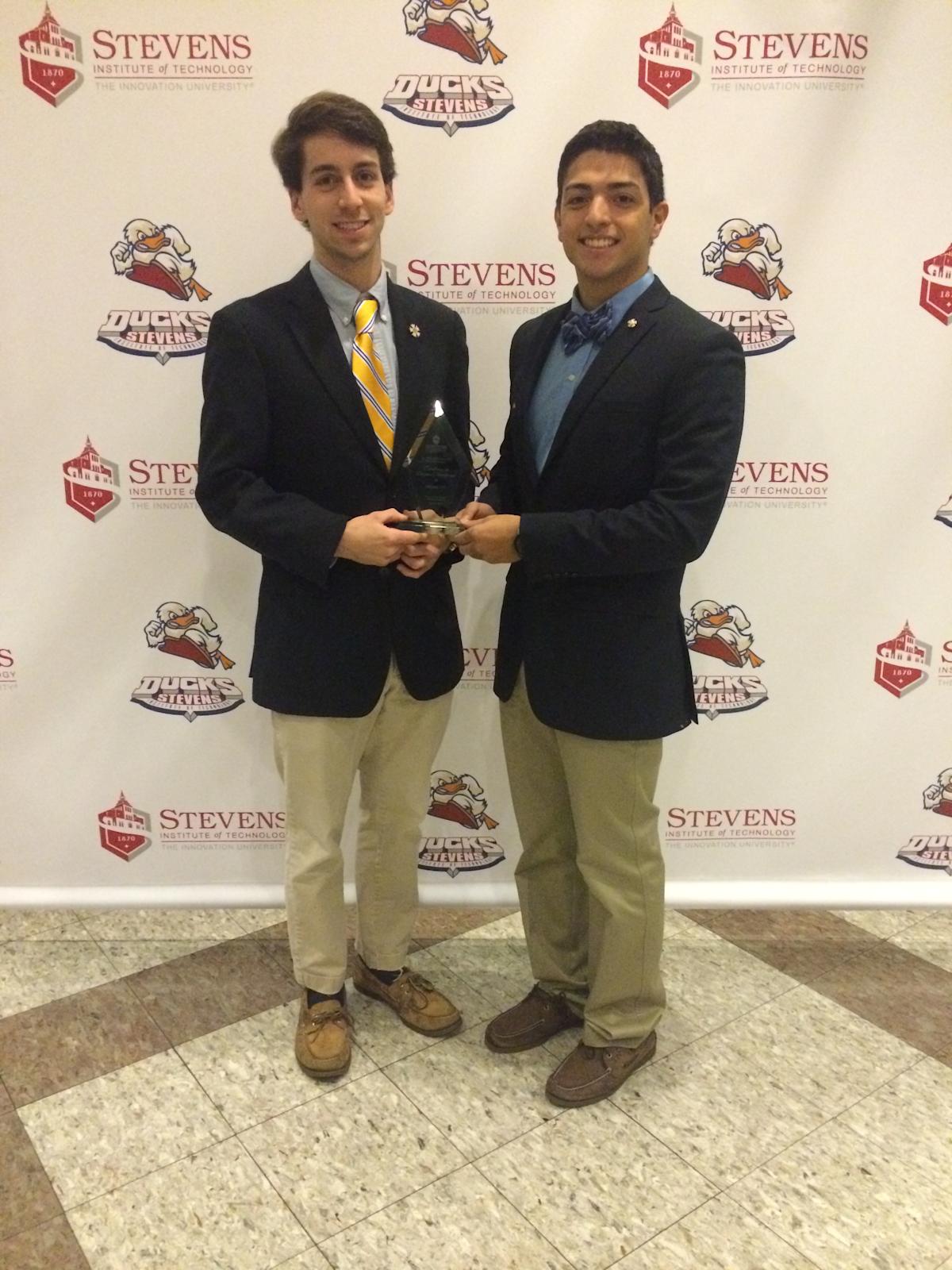 Two young men in navy blazers and khaki pants stand together holding a glass award trophy, smiling in front of a repeating step-and-repeat backdrop featuring a university's name and duck mascot logo.