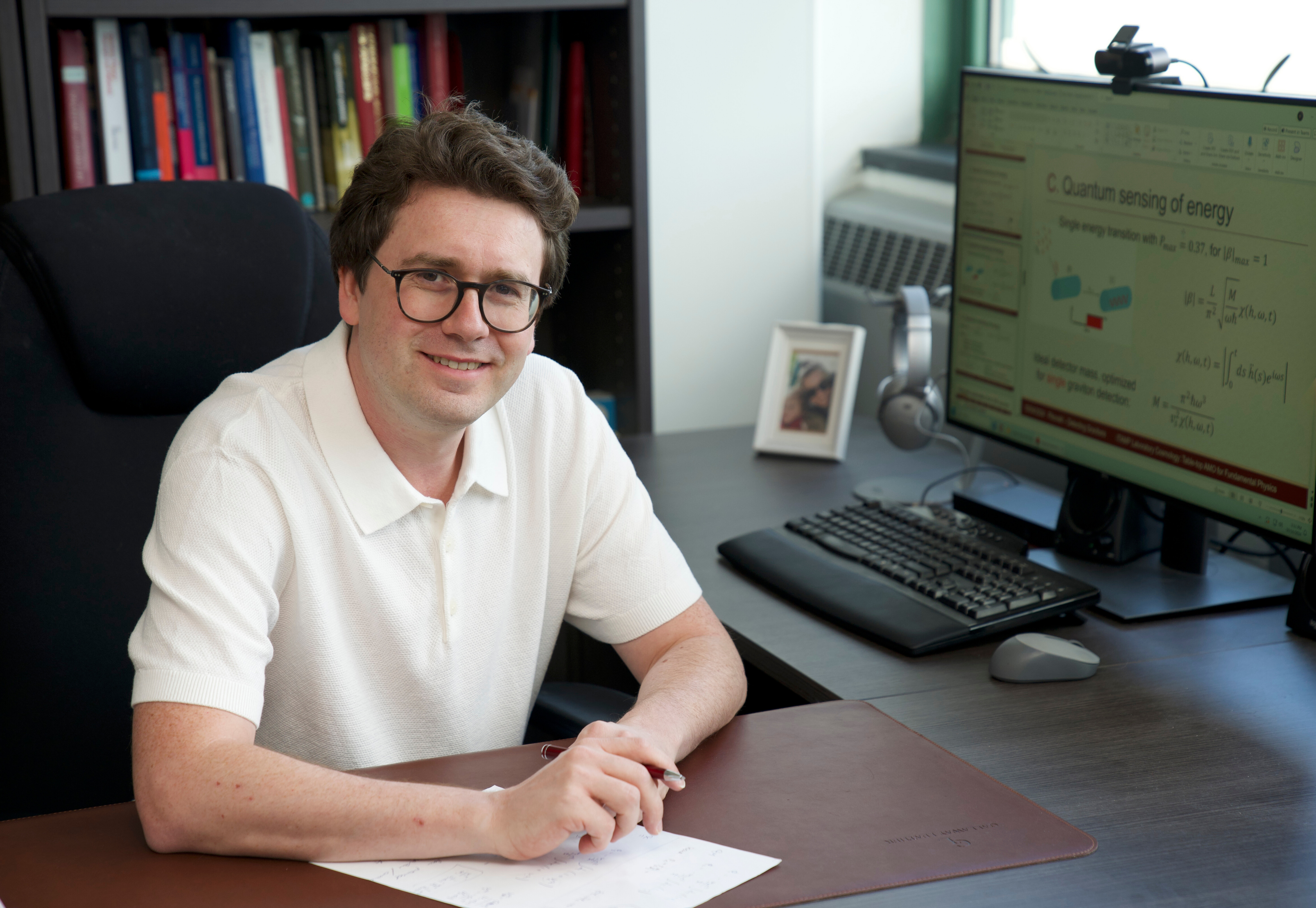 Stevens professor Igor Pikovski faces the camera with a pen in hand and paper on desk with a computer monitor in the background.