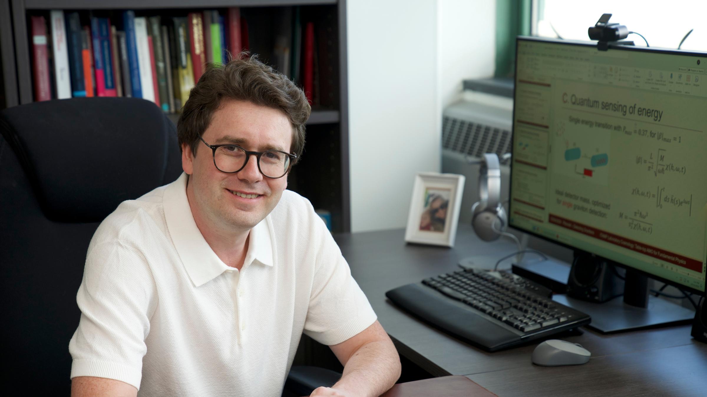 Stevens professor Igor Pikovski faces the camera with a pen in hand and paper on desk with a computer monitor in the background.