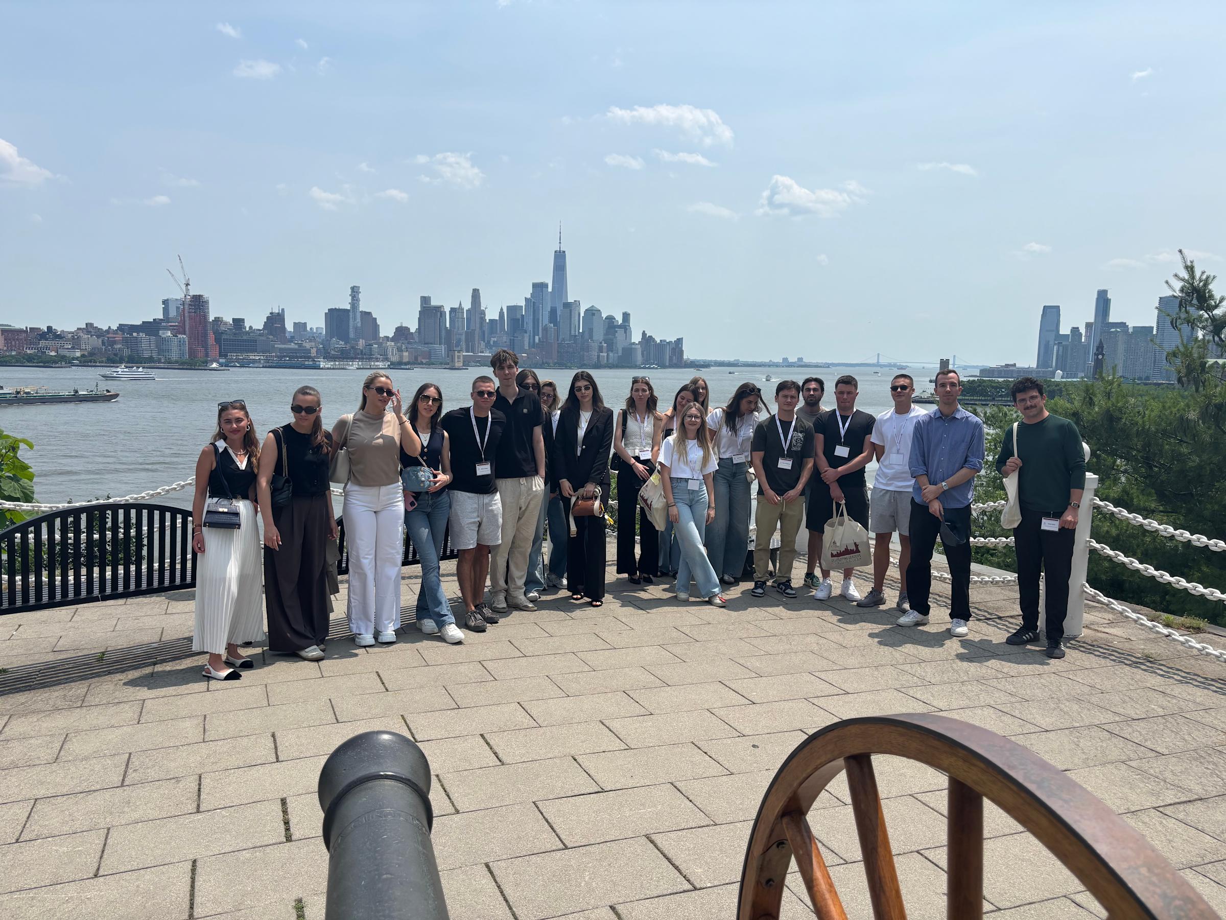 A large group of students wearing conference badges stands on a waterfront plaza with a historic cannon in the foreground and the Manhattan skyline featuring One World Trade Center across the harbor on a sunny day.