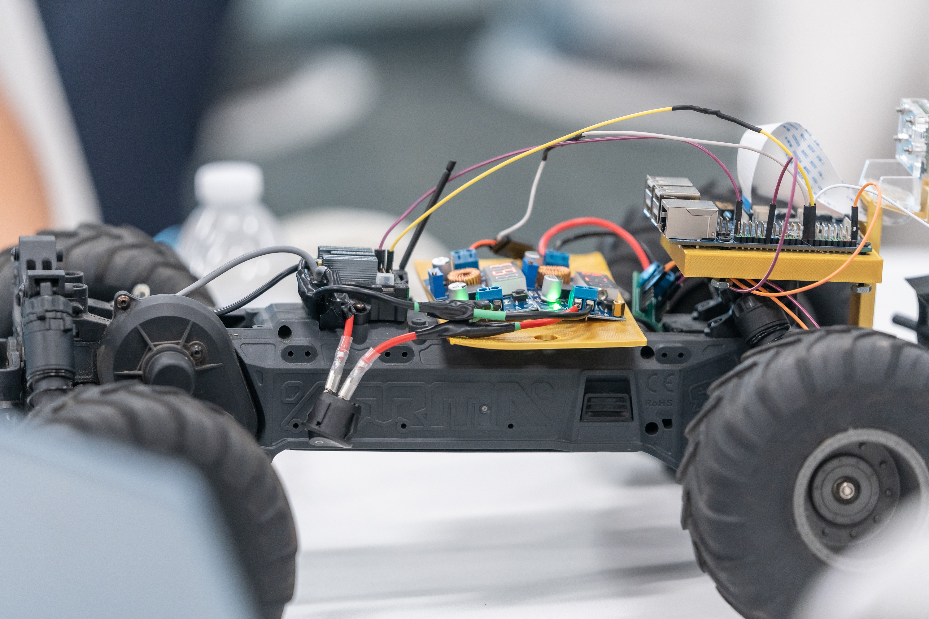 Close-up of an ARRMA RC truck chassis converted into a robot car, with a Raspberry Pi on a gold 3D-printed mount, two voltage converter boards with red LED displays, a camera ribbon cable, and colorful jumper wires.