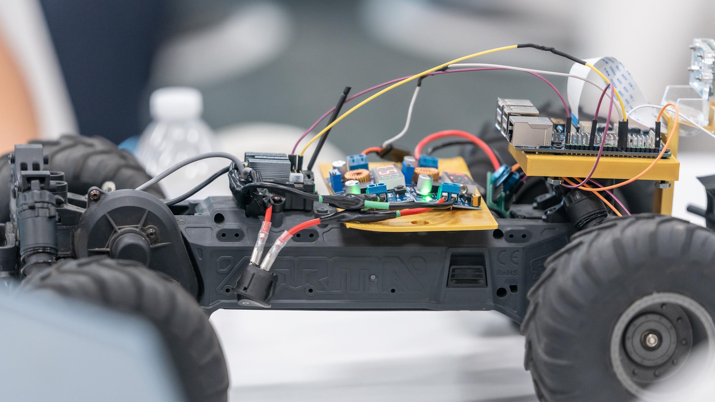 Close-up of an ARRMA RC truck chassis converted into a robot car, with a Raspberry Pi on a gold 3D-printed mount, two voltage converter boards with red LED displays, a camera ribbon cable, and colorful jumper wires.