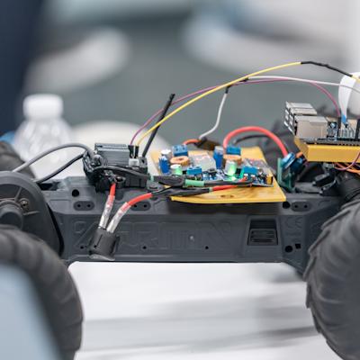 Close-up of an ARRMA RC truck chassis converted into a robot car, with a Raspberry Pi on a gold 3D-printed mount, two voltage converter boards with red LED displays, a camera ribbon cable, and colorful jumper wires.