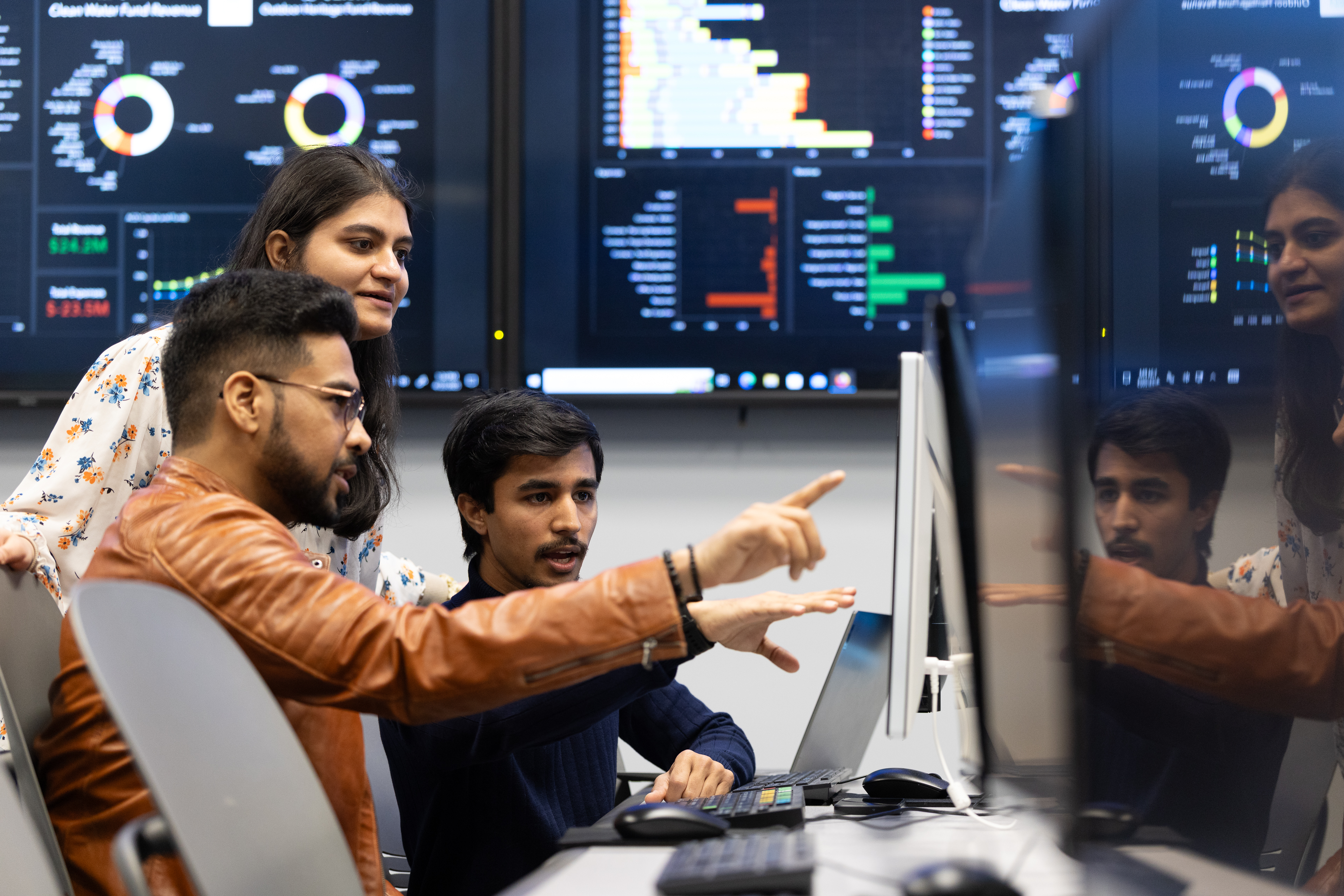 Three students collaborate at a computer in a financial systems lab, with large wall-mounted monitors displaying data visualizations, charts, and analytics in the background. One student points at the screen while explaining something to the others.