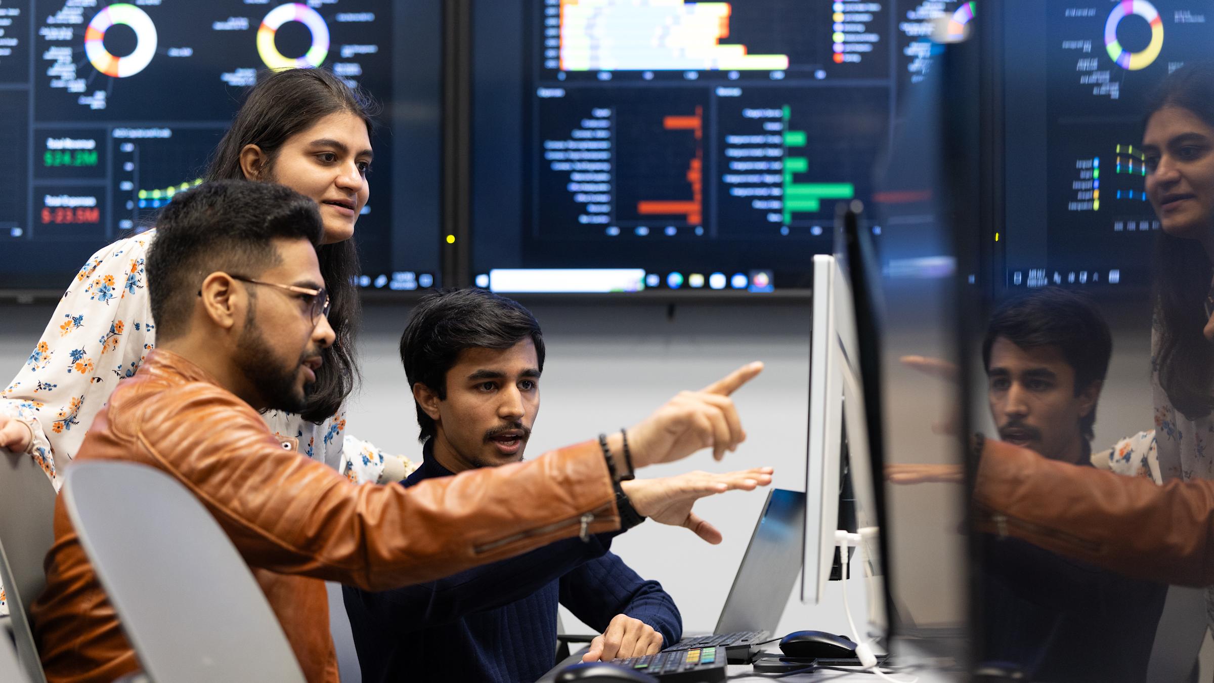 Three students collaborate at a computer in a financial systems lab, with large wall-mounted monitors displaying data visualizations, charts, and analytics in the background. One student points at the screen while explaining something to the others.