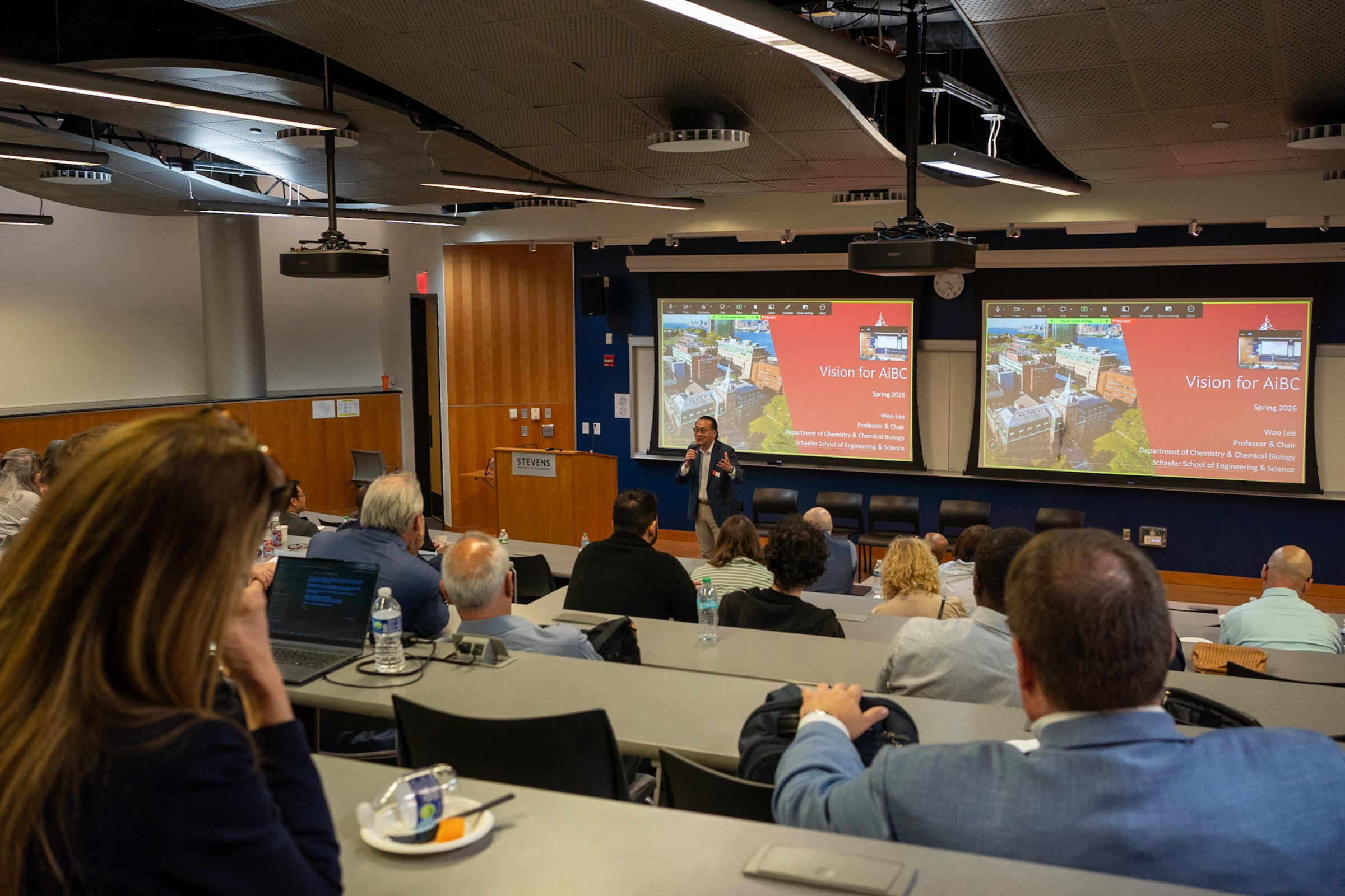 Woo Lee, the leader of the Community for Artificial Intelligence in Biology and Chemistry stands at the front of a lecture hall giving a presentation to community members, students and educators.
