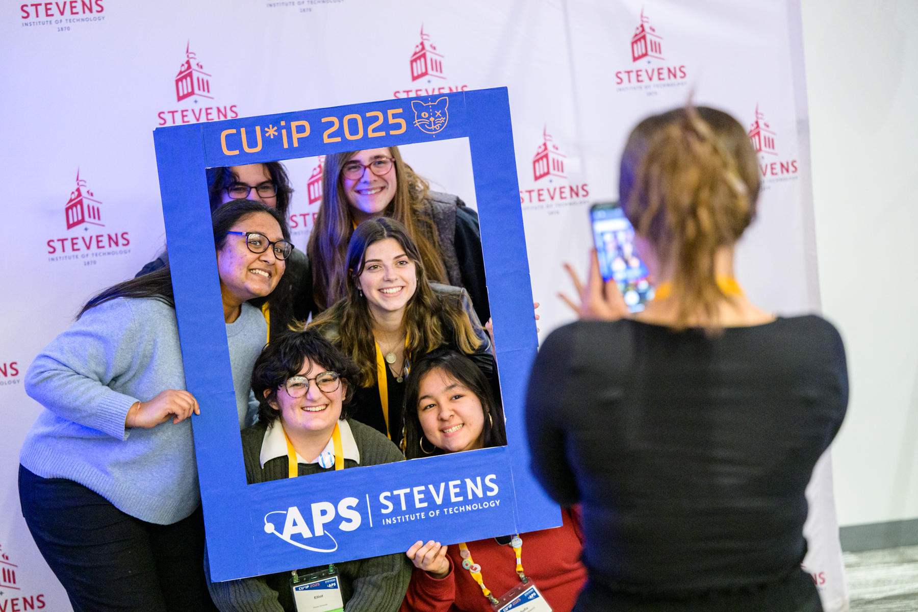 A group of students pose for a picture behind a branded large frame using Stevens, American Physics Society and CUWIP branding.
