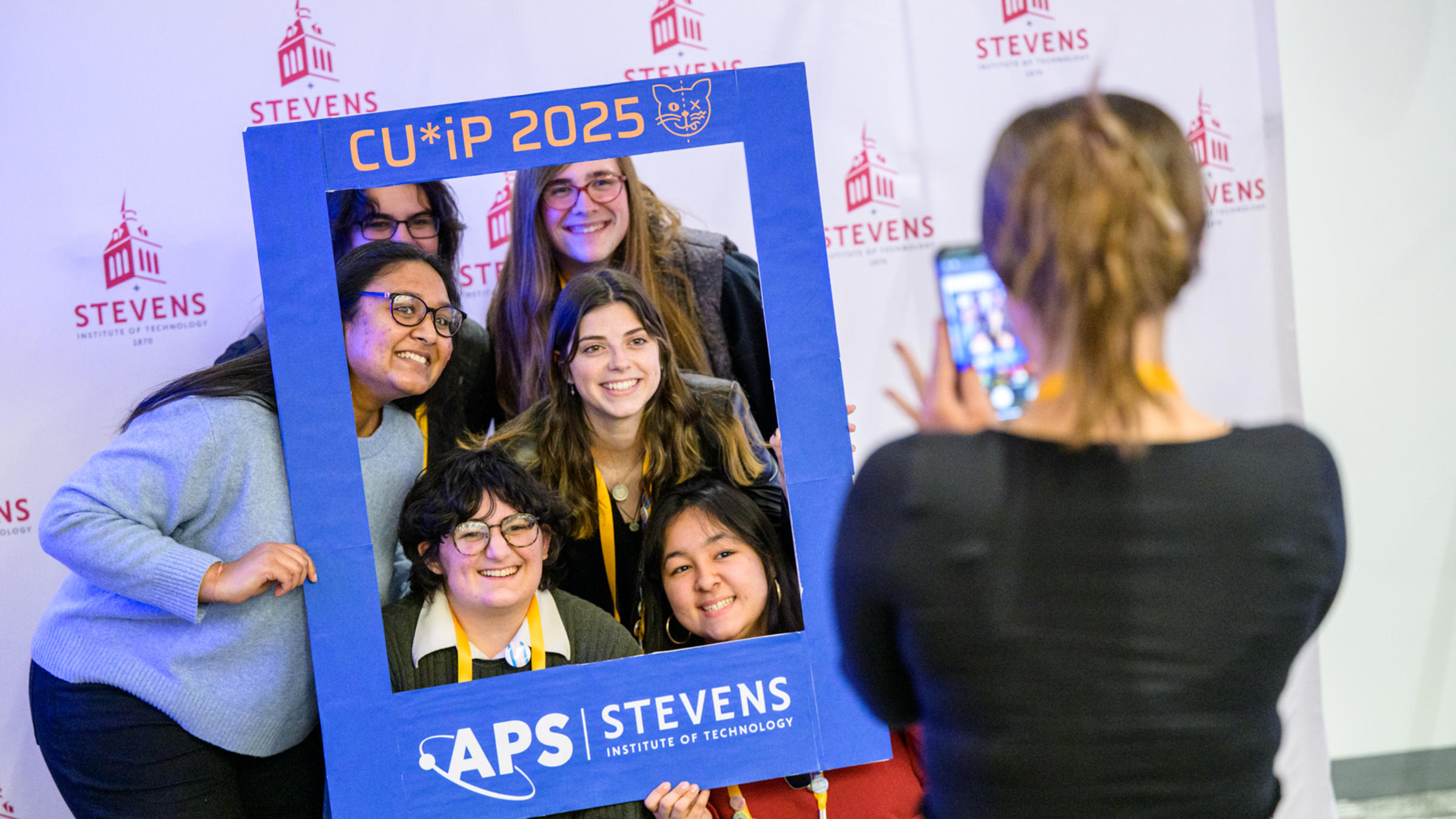 A group of students pose for a picture behind a branded large frame using Stevens, American Physics Society and CUWIP branding.