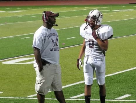 A football player in a white uniform with helmet in hand stands beside a coach wearing a maroon cap and white shirt on a turf field at night, with spectators lining the track in the background.