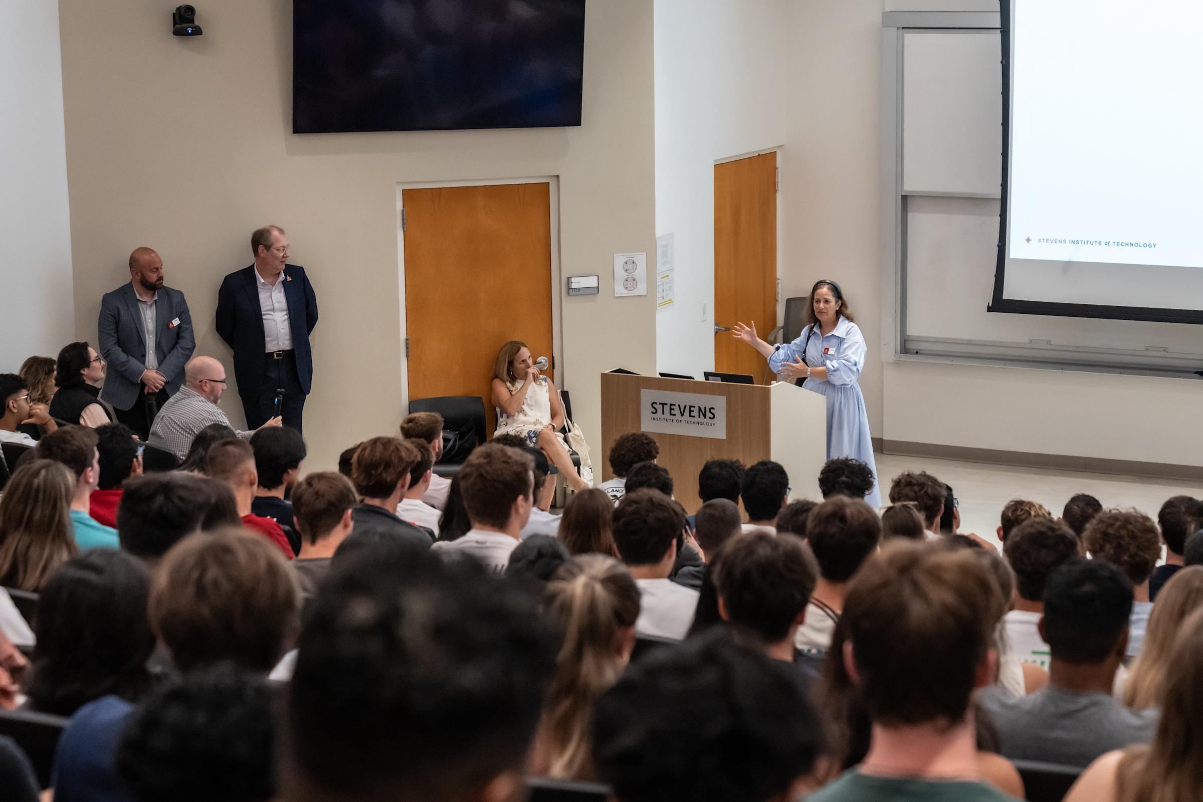 Female speaker presenting at Stevens Institute podium to packed student audience, with faculty panel seated along wall in modern classroom.
