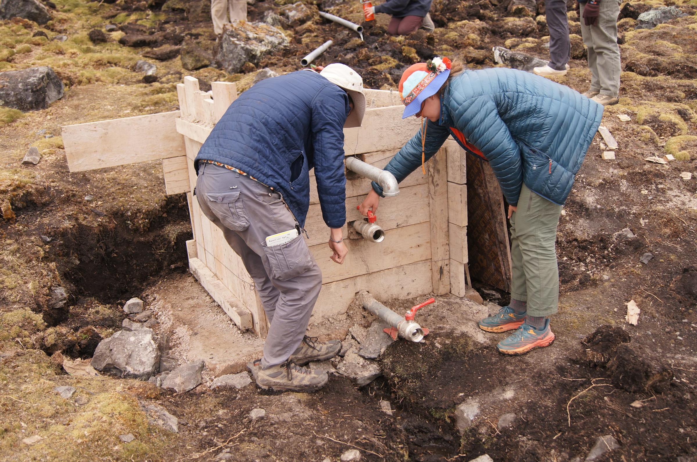 Two people crouch over a wooden box with drain pipes sticking out of it.
