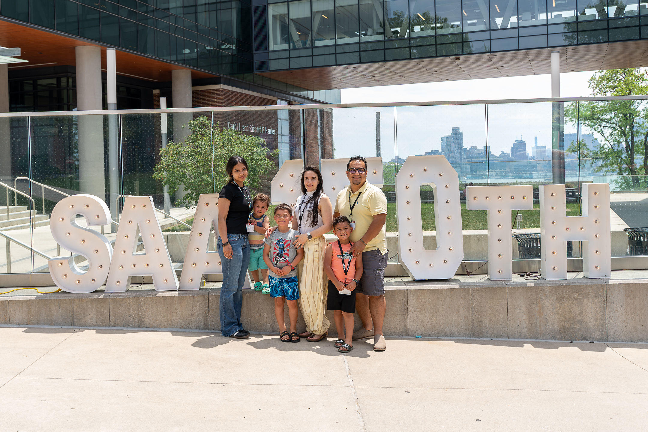 A family of six posing outdoors in front of an SAA 150th sign at Alumni Weekend 2026