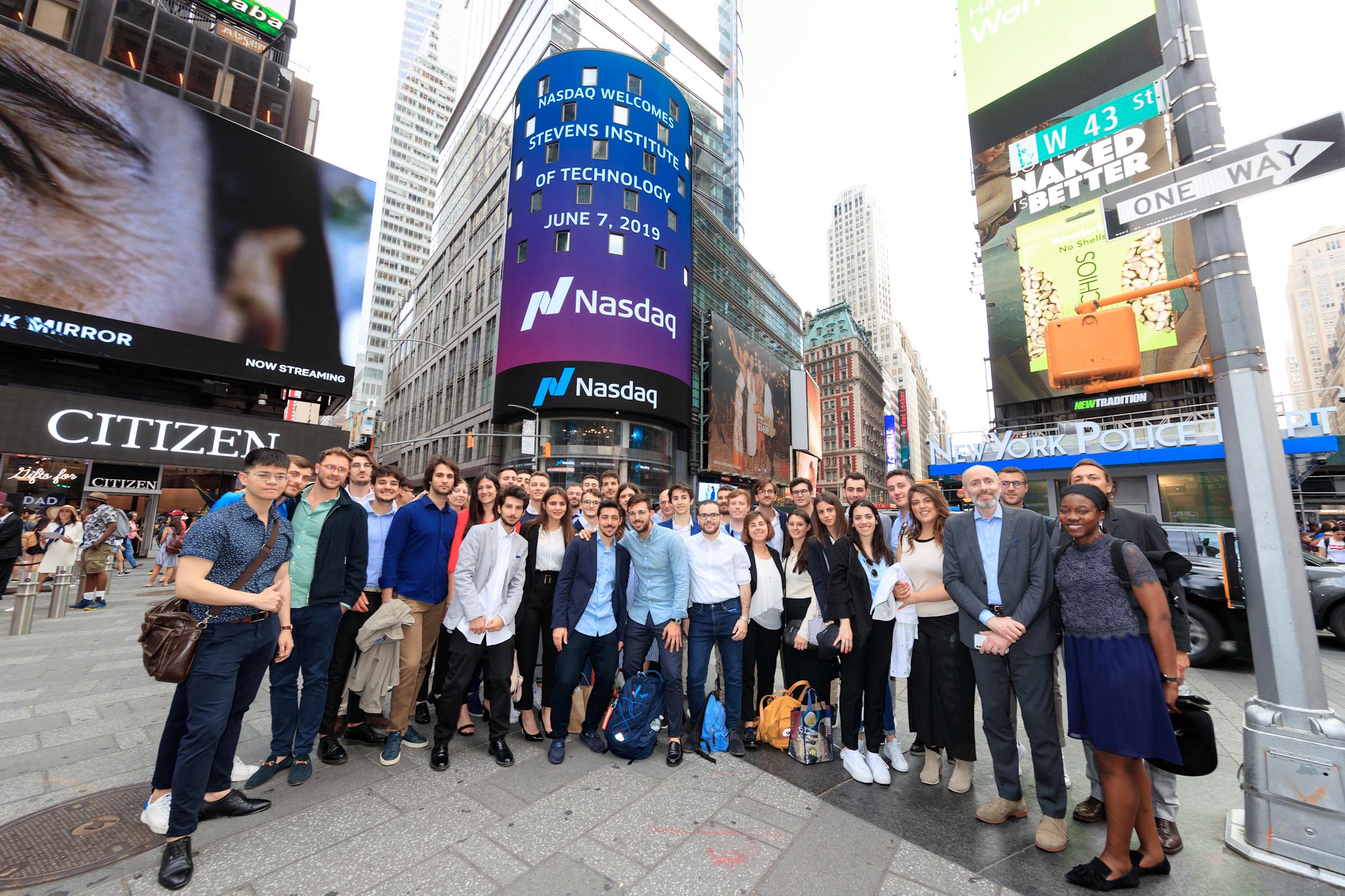 Large group of Stevens Institute students and faculty pose in Times Square beneath a Nasdaq tower displaying "Nasdaq Welcomes Stevens Institute of Technology — June 7, 2019" with W 43 St street signs visible overhead.