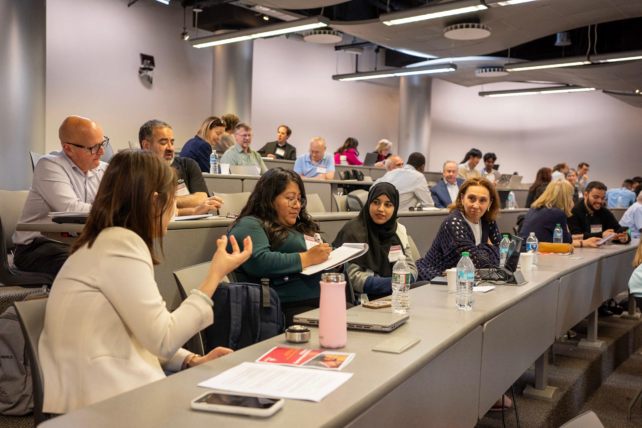 A row of four women sitting in the same row in a lecture hall having a conversation and making hand gestures while being engaged in conversation with each other.